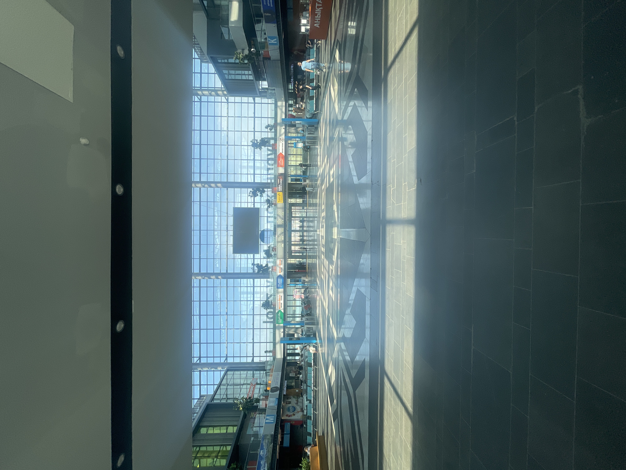 The image shows the interior of a modern, spacious airport terminal with a high, glass-paneled ceiling and walls, allowing natural light to flood the area. The floor is tiled with large, dark gray tiles, and there is a patterned design of lighter gray tiles in the central area, creating a reflective surface.
In the middle ground, there are several rows of teal-colored seating arranged for waiting passengers, with some individuals seated. Beyond the seating, automatic glass doors lead to an outdoor area or another part of the terminal. A large digital display screen is visible mounted high on the wall behind the glass facade. Various signs and advertisements are present on the walls and pillars, some in what appears to be Cyrillic script.
On the right side of the terminal, a person in a blue t-shirt and jeans walks towards the viewer, while another person in a black t-shirt walks away. Further to the right, a counter with a sign that reads "АНЫҚТА" (which means "confirm" or "identify" in Kazakh) is staffed by two people.
The overall environment suggests a clean, well-lit, and busy transportation hub. Given the presence of signs in Cyrillic script and the mention of Astana, Kazakhstan, this is likely the Nursultan Nazarbayev International Airport (NQZ) in Astana, Kazakhstan. The lighting indicates it is daytime, with sunlight casting distinct shadows across the floor. The architecture is contemporary, featuring a lot of glass and geometric lines.