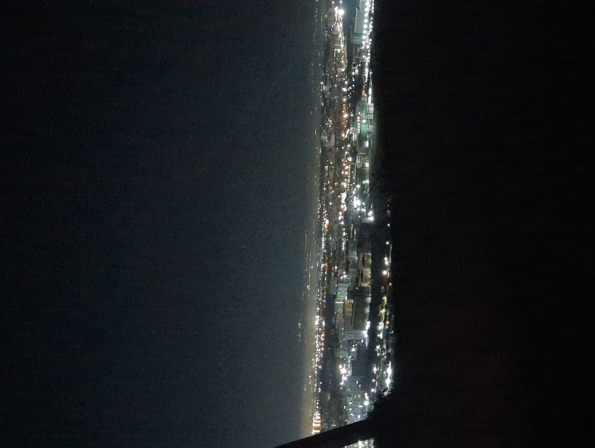 The image shows a night view of Bishkek City, Kyrgyzstan, characterized by city lights stretching across the scene. The left side of the image consists of a dark expanse, possibly a body of water or open land. The right side presents clusters of illuminated buildings and streets, forming the cityscape. The photograph seems to be taken from a high vantage point, possibly an elevated building or a natural height. The light from buildings and streetlights are visible, creating a contrast against the dark background. No people are visible in the image.