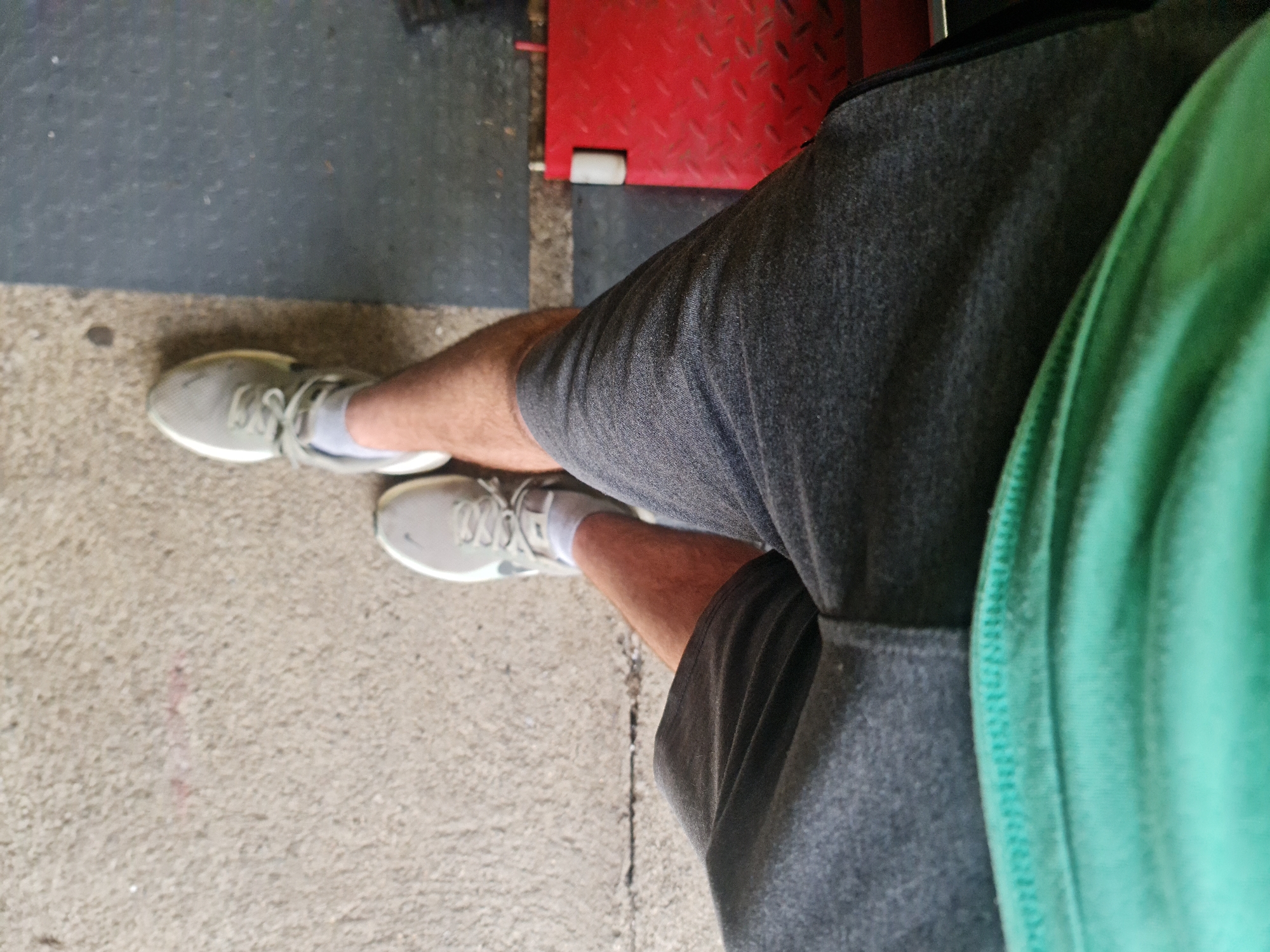 The image shows a first-person, downward-looking perspective of a person's legs and lower torso. The individual is wearing gray athletic shorts and a sliver of a green shirt is visible on the right. Their legs are bare and appear to be tanned. They wear white ankle socks and light gray athletic shoes with white soles and matching gray laces. A dark, possibly black, logo mark is faintly visible on the side of the shoe closest to the viewer.
The flooring consists of two distinct sections: a dark gray, possibly rubber or textured mat, covers the upper left portion and extends to the left, while a lighter, rougher concrete or asphalt surface occupies the lower right and bottom. A clear seam or transition line runs diagonally across the image, separating these two surfaces. In the upper right background, a bright red, diamond-plate textured metal surface is visible, suggesting industrial or workshop equipment. A small, white cylindrical object rests near the red surface. The lighting suggests it is daytime. The scene is static, with the person either standing or sitting. The location is Buzovgrad, Bulgaria.