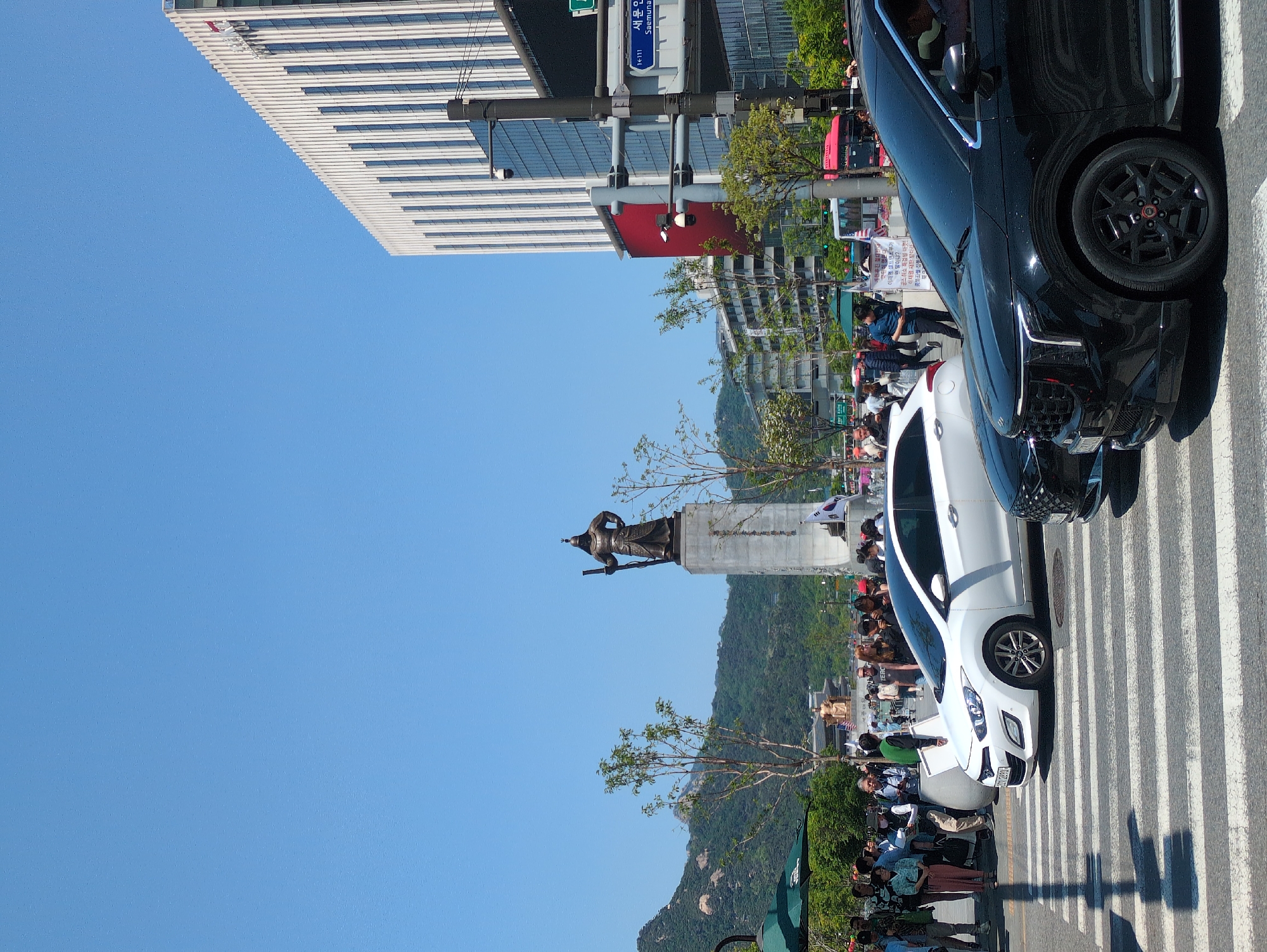 The image captures a street scene in Seoul, South Korea. The statue of Admiral Yi Sun-sin is visible in the background. A crowd of people is gathered near the statue. The foreground contains two parked cars, a white hatchback and a black SUV, positioned on a zebra crossing. A tall office building with a striped facade is seen in the upper left corner. A street sign with Korean and English text is mounted on a pole near the office building. A mountain is located in the background beyond the statue.