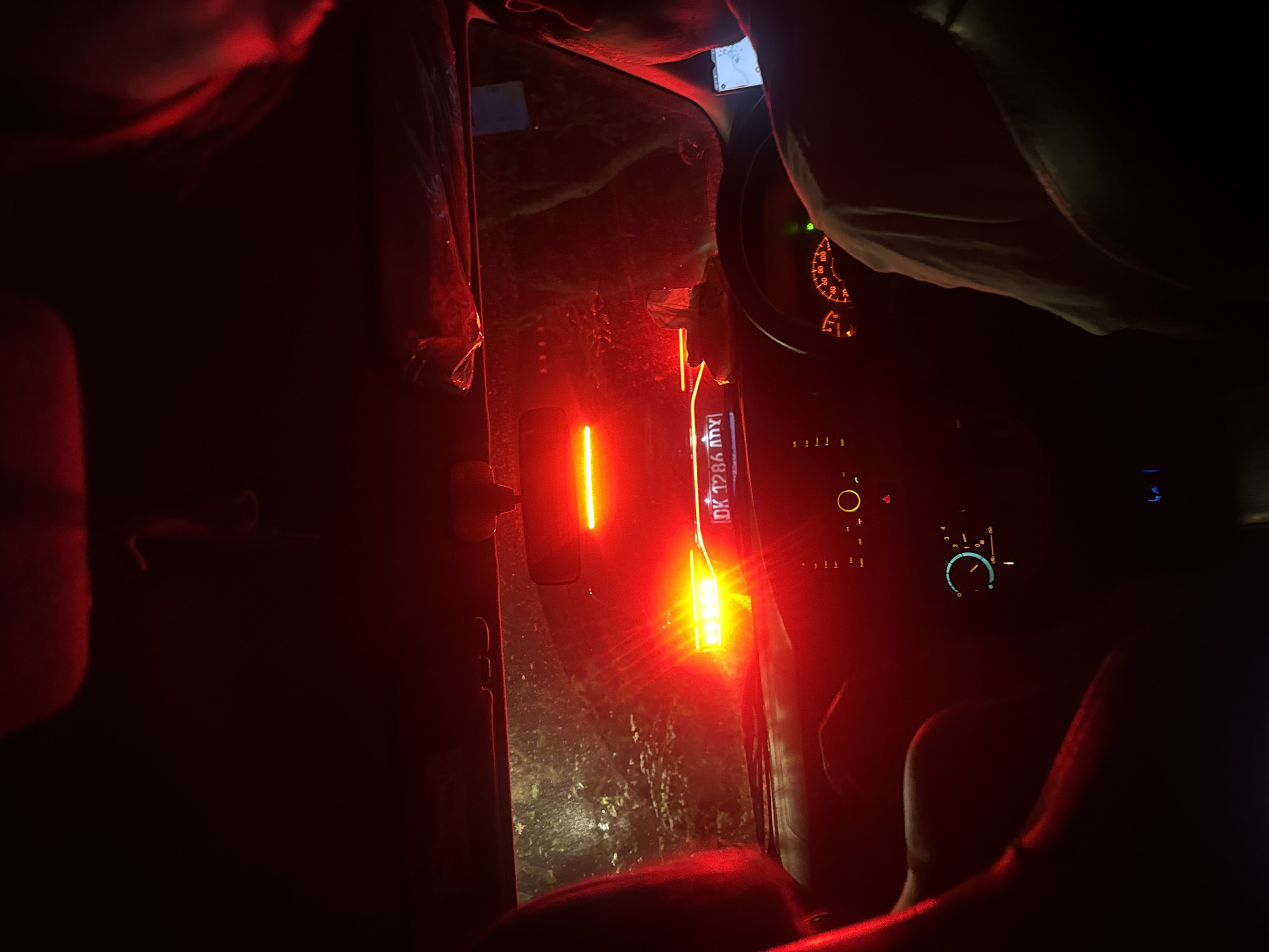 The image, heavily dominated by red illumination, shows the interior of a car at night, with the apparent location being Kuta Selatan, Indonesia, as inferred from the obscured number plate "DK 128A my". A person is seen from an overhead perspective, their arm partially visible at the top right, and possibly another person's shoulder on the left. The dashboard and steering wheel of the car are visible, illuminated by the instrument panel, which includes a speedometer. A rectangular light source, likely an emergency signal or reflective panel, is situated on the passenger side of the windshield, with additional orange and red lights shining outwards. The environment outside the car appears dark and textured, possibly indicating a road or vegetation. Car seats, partially visible, are draped in shadow.