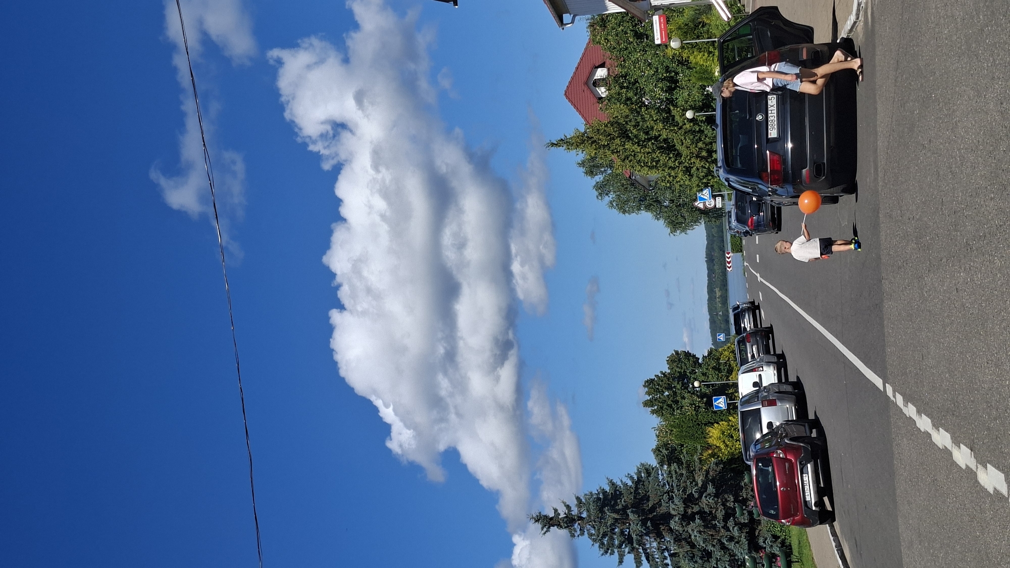 The image depicts a street scene in Myadzyel, Belarus, on a sunny day with a bright blue sky and scattered white clouds. A black SUV is parked on the right side of the road with its passenger door open. A young boy wearing a white t-shirt and dark shorts is standing next to the SUV, holding an orange balloon on a string. A woman, also wearing a white t-shirt and denim shorts, is standing near the open SUV door, appearing to interact with the boy. Several other cars are parked along the left side of the road, which curves towards a body of water visible in the distance. The left side of the street features a grassy area and tall evergreen trees, while the right side has houses and trees. Traffic signs are visible on the left side of the road.