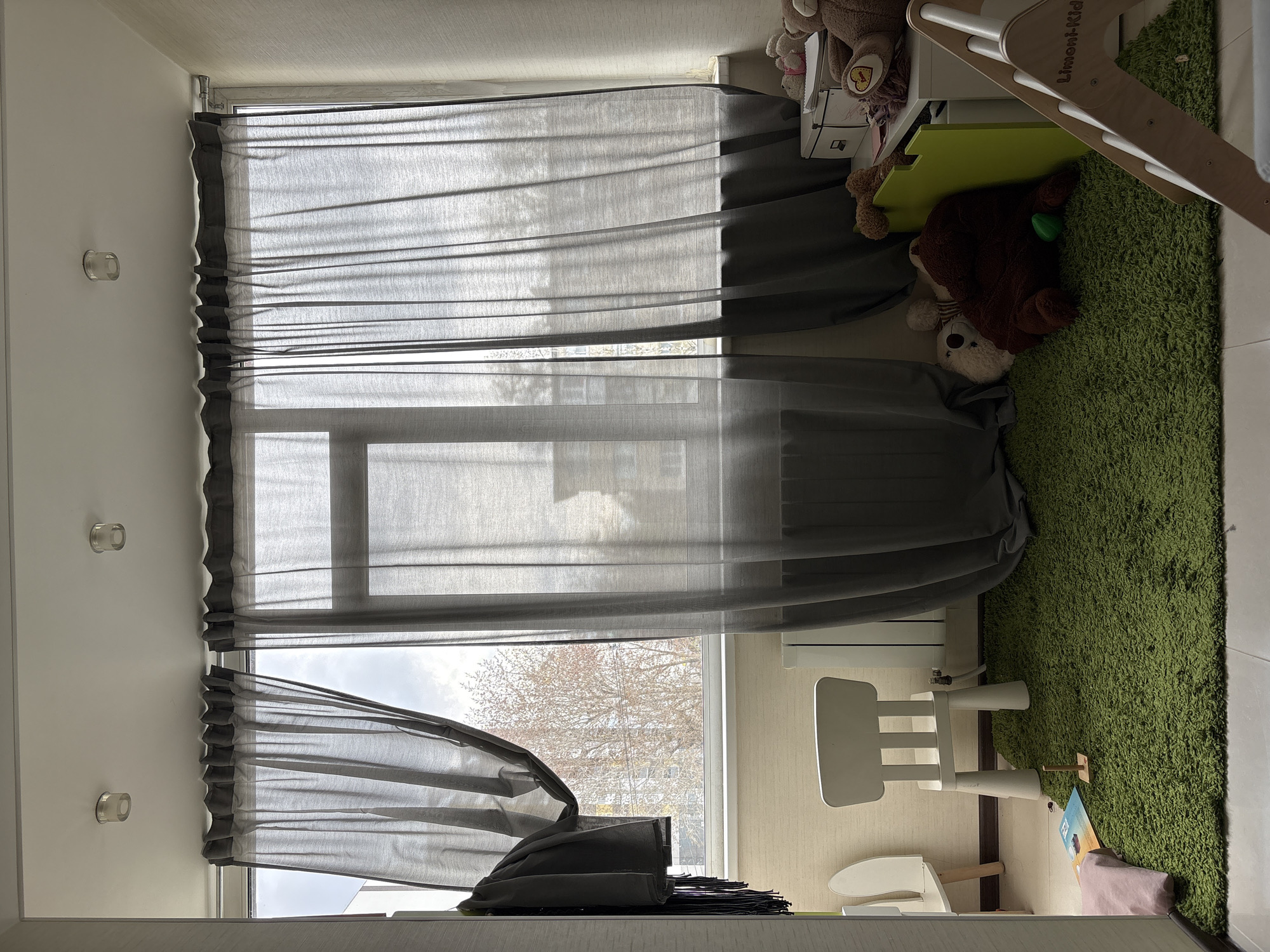 A child's room in Ulyanovsk, Russia, features a window covered with semi-sheer gray curtains. The lower section of the curtain is partially open, revealing a view of a bare tree and cloudy sky outside. Inside the room, a green shag rug covers part of the floor. A white child's chair and a small, similar-style seat are visible on the rug, near a radiator. Several stuffed animals are placed near the window. A child's play structure is partially visible.