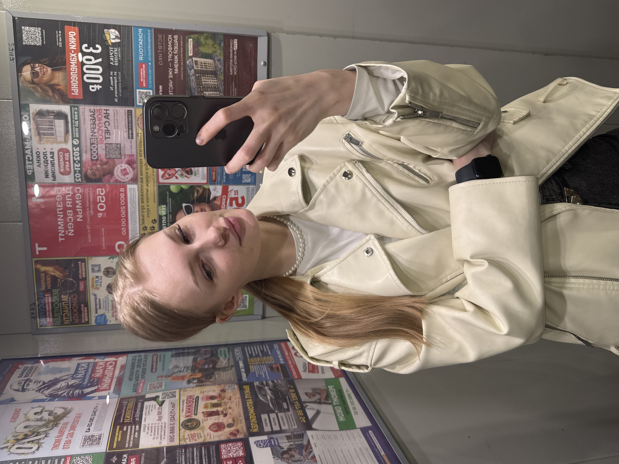 A young woman is captured in a selfie, holding a dark-colored smartphone with her right hand, its multi-lens camera visible. She has light-colored hair and a neutral expression, looking towards the phone's screen. She is wearing a light-colored, possibly cream or off-white, faux leather jacket with multiple zippers and snap buttons. A white T-shirt and a pearl necklace are visible beneath the jacket. A dark smartwatch is worn on her left wrist.
The setting appears to be indoors, against a large display board densely covered with various printed advertisements and flyers, mostly in Russian. The image orientation is rotated ninety degrees clockwise. The background is a collage of text, images, and QR codes, advertising diverse services and products. Notable visible text includes prices such as "3 000 ₽" and "220 ₽", along with phone numbers like "8 800 520 00 20" and "503-21-05". Several Russian words and phrases are discernible, such as "СКИДКИ" (Discounts), "НЕДВИЖИМОСТЬ" (Real Estate), "КОМФОРТ" (Comfort), "АКЦИЯ" (Promotion/Sale), and "ОКНА" (Windows). The presence of numerous advertisements suggests a public area, possibly in Кондратово, Russia, as per the location context. The lighting suggests an indoor environment.