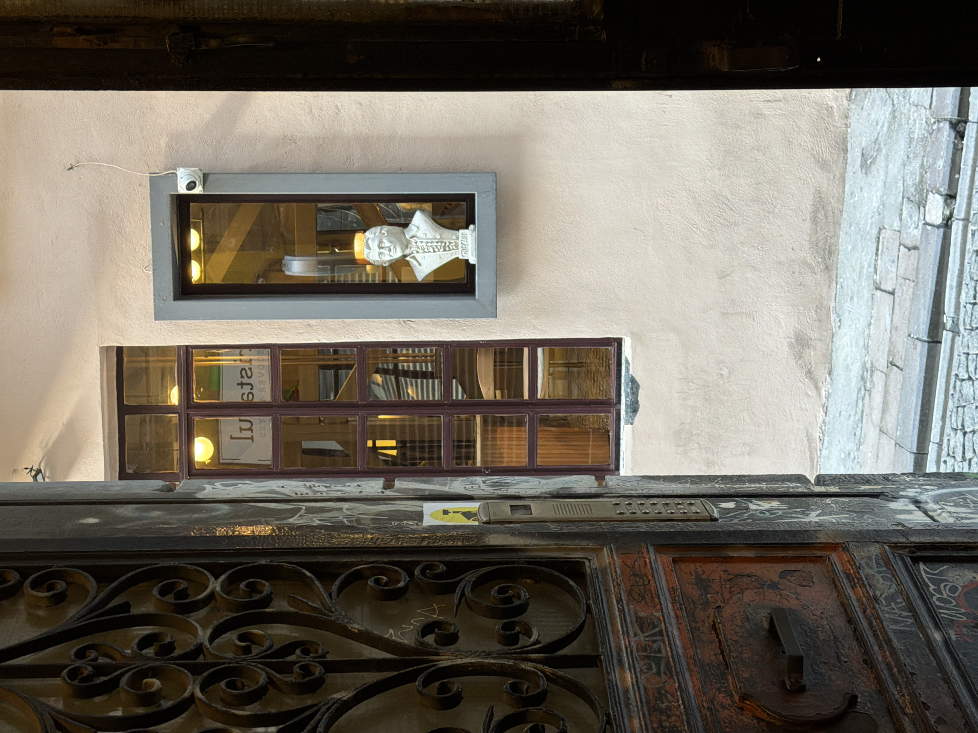 The image shows an outdoor scene in the Beyoğlu district of Istanbul, Turkey, focusing on the entrance to a building and its facade. In the foreground to the left, a weathered wooden door with ornate metalwork, possibly a gate or part of a larger entrance, is ajar, revealing graffiti on its surface. To the right of the door, a light-colored plastered wall dominates the scene. A rectangular window with a dark frame, divided into panes, is visible on the wall. Through the window, reflections and interior lighting suggest a space within the building. Slightly above and to the right of this window, another smaller, taller window is set into a gray frame. Inside this upper window, a white bust of a person, possibly historical, is displayed. A small white security camera is mounted on the wall near the top right corner of this framed window. The ground in front of the building is paved with cobblestones.