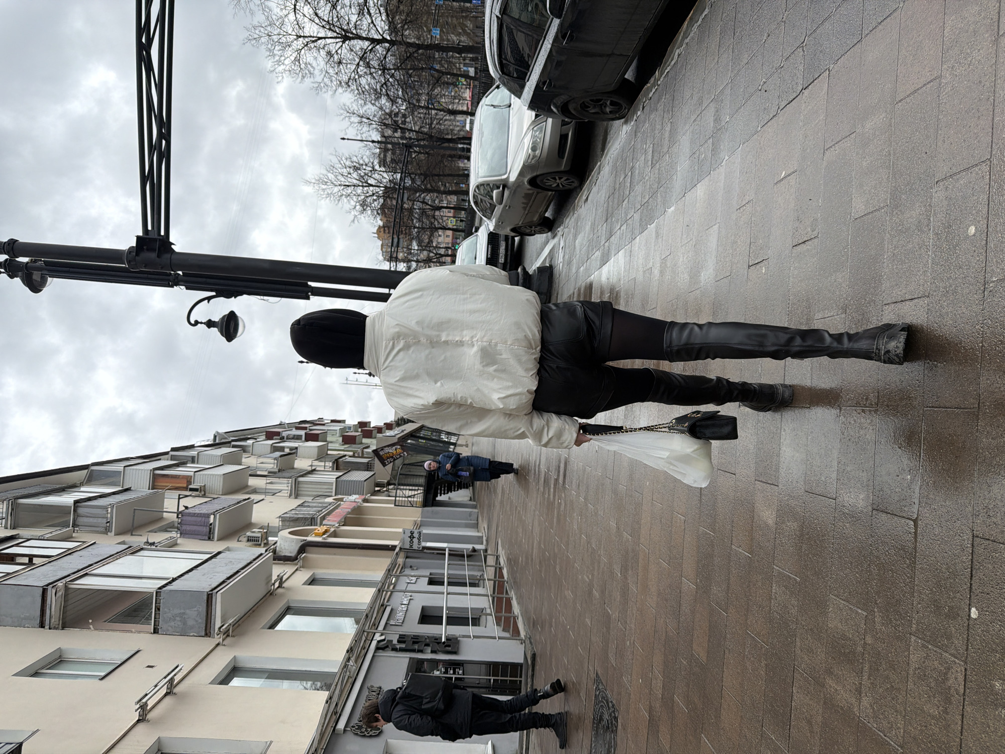 A woman wearing a white jacket and black leather-like pants and boots walks on a brick sidewalk on a cloudy day. She carries a white plastic bag and a black purse. A black lamppost is on the left. Parked cars line the street to the right. Other pedestrians are walking further down the sidewalk. Buildings with balconies and window boxes line the street on the left. A man wearing a black jacket walks on the sidewalk in the opposite direction. The location is Perm, Russia.