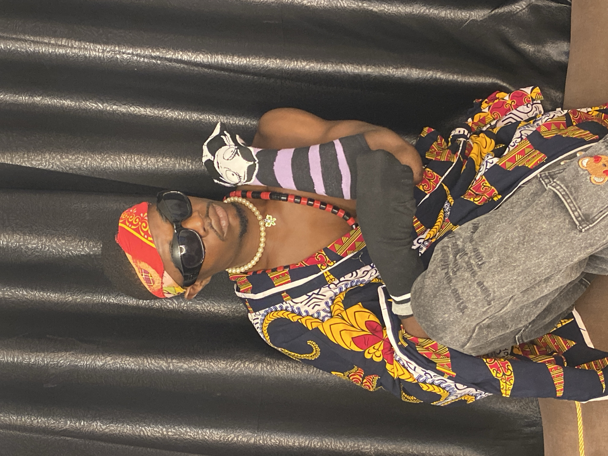 A person is posed in front of a black, textured curtain. The person is wearing sunglasses, a red bandana with a yellow and white pattern, a pearl necklace, and a red beaded necklace with a pendant. They are also wearing a colorful, patterned robe with yellow, red, blue, and white designs. The robe is open, revealing their chest. The person is holding a sock with a cartoon character's face on it and purple and black stripes. They are also wearing grey jeans with a teddy bear design on the pocket. The person is kneeling and resting their elbow on their knee. The scene does not provide any specific information to determine a location within the city of Abakaliki, Nigeria.