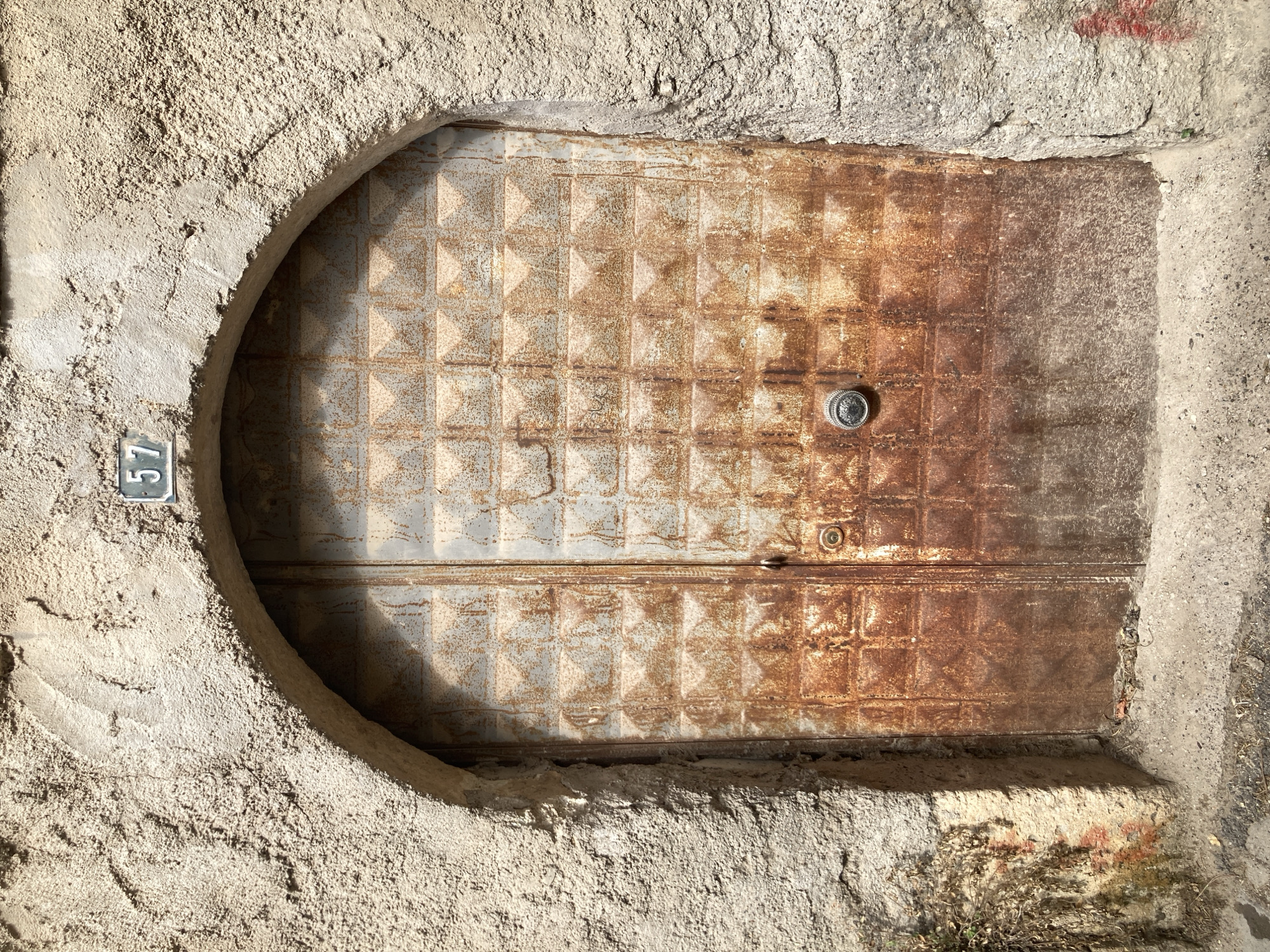 A heavily rusted, double metal door is set within a rough, arched stone entryway. Above the arch, a small rectangular sign displays the number "57". The door's surface is patterned with a grid of raised squares, each containing a pyramidal indent. The left side of the door appears to have a vertical seam, suggesting it might be a double door or a large single door with a visible frame detail. A circular doorknob, also metallic and showing signs of age and wear, is located on the right side of the door. The stone walls surrounding the doorway are textured and weathered, with some areas showing evidence of moss or vegetation growth and faint red markings. The ground in front of the door is paved with concrete or similar material, with some debris scattered on it. The scene is outdoors, in what appears to be a somewhat aged urban environment.