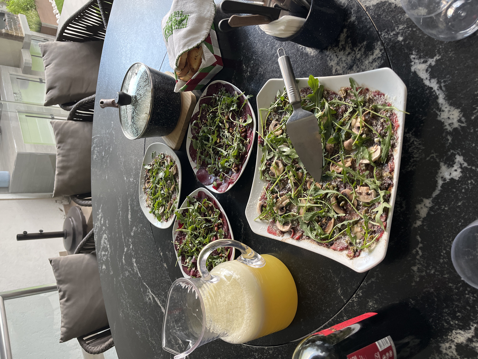 A table is set for a meal outdoors. There are four plates, one square and three oval, with what appears to be arugula and sliced meat on top. A black pot with a glass lid and wooden handle is on the left side of the table. Next to the pot is a white cloth with green embroidered detailing, partially covering a bowl of bread. Silverware is in a black container. A glass pitcher with yellow liquid is towards the bottom of the frame, next to a bottle of red wine. A wine glass sits at the edge of the table. The table has a dark, marbled surface and is surrounded by chairs with gray cushions. The location is San Bernardino Tlaxcalancingo, Mexico.