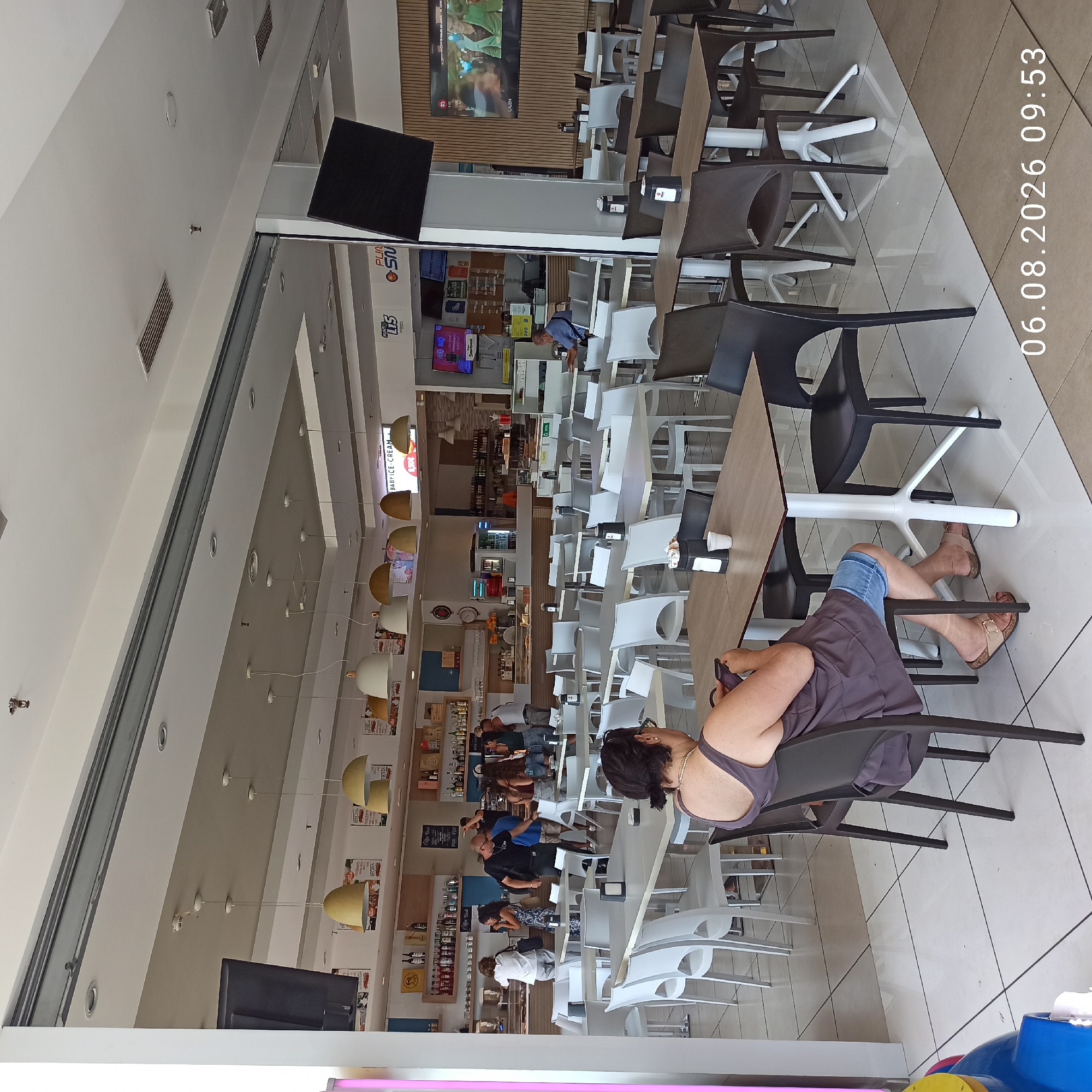 The image depicts the interior of a cafe or restaurant. A woman wearing a dark grey tank top and blue denim shorts is seated at a table, looking at her phone. She is wearing brown sandals. The table has a black napkin holder and a white cup on it. There are several other tables and chairs in the background, some occupied by other patrons. The cafe appears to be brightly lit with natural light from large open doorways and artificial lighting from pendant lamps. There are shelves with bottles and merchandise visible behind the counter. The floor is tiled with a checkered pattern. A digital clock overlay in the bottom right corner reads "06.08.2026 09:53". The location is identified as Bari, Italy.