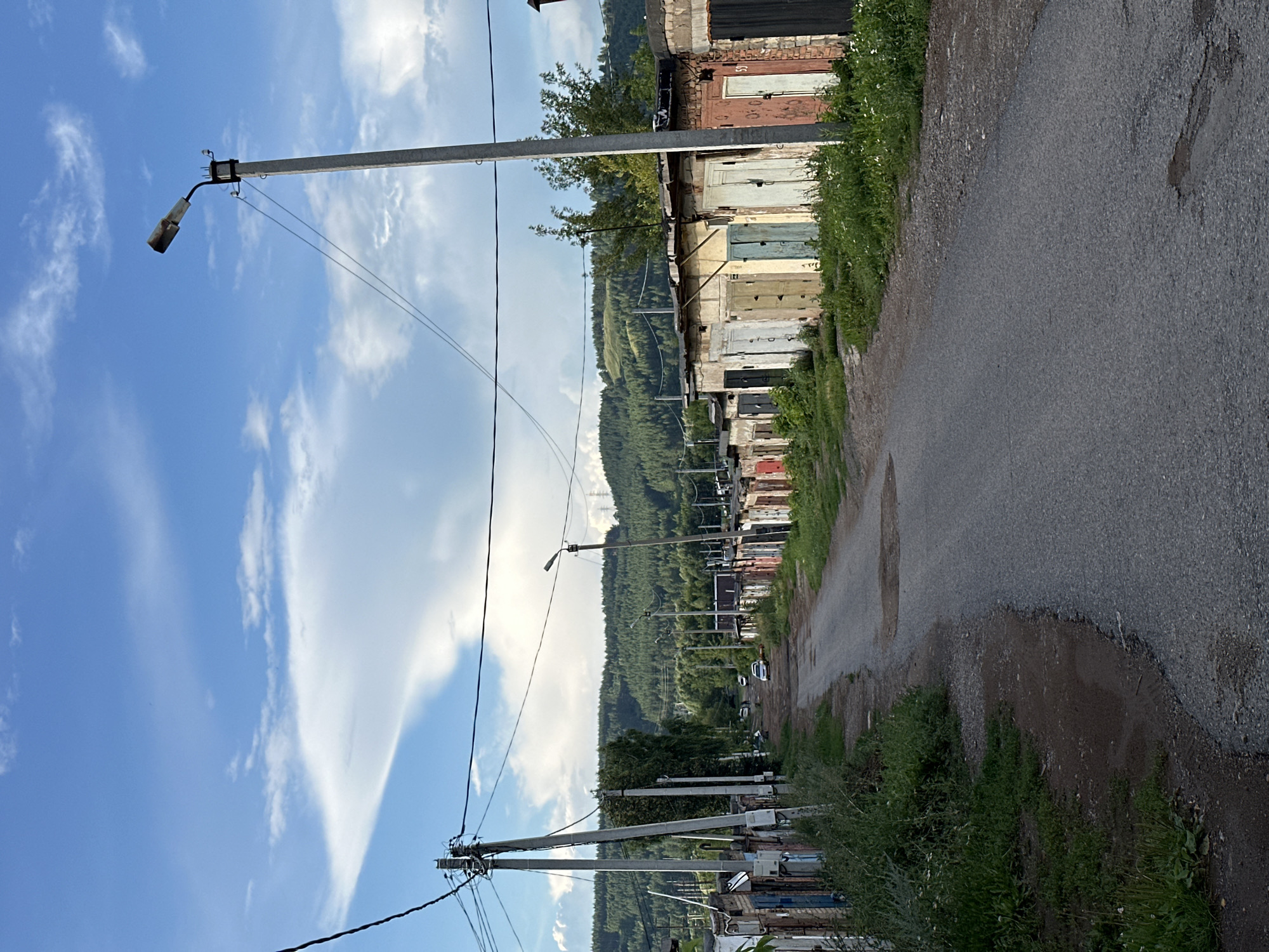 The image depicts a row of garages lining a paved road in a suburban or semi-rural area. The garages appear to be constructed from brick and metal, with various colored doors. Green grass and vegetation grow around the garages and along the roadside. In the background, a dense forest covers rolling hills, under a blue sky with scattered white clouds. Several cars are visible parked along the road. Power lines and utility poles are present throughout the scene, indicating infrastructure. The overall impression is one of a functional, lived-in environment.
