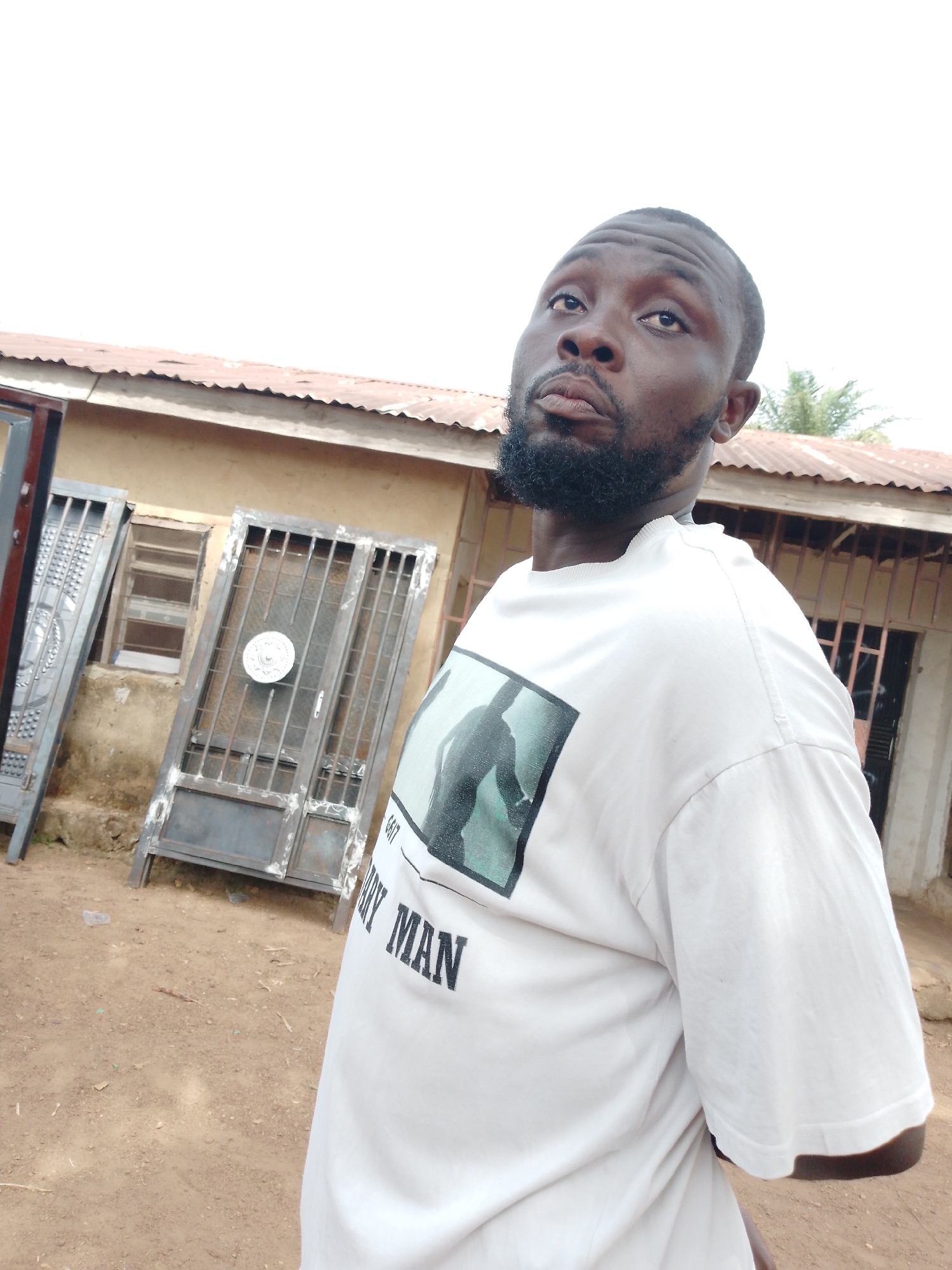 A medium shot captures a man posing outdoors, looking upwards and slightly to his right with a serious, contemplative expression and slightly pursed lips. He has a dark, full beard and short hair. He is wearing a loose-fitting, white short-sleeved t-shirt. The t-shirt features a prominent black graphic print on the front, including a rectangular image that appears to be a dark silhouette. Below this image, partial text is visible, reading "RY MAN", and above it, possibly "6617".
The background shows a single-story building with a light-colored, possibly beige or yellow, wall and a rusty, corrugated metal roof. In front of the building, several large metal gates or doors are visible, some standing upright and others partially obscured. One prominent gate in the center has vertical bars and a circular decorative emblem. A small window is also visible on the building's facade. The ground is unpaved, consisting of bare earth or dirt. The sky above is overexposed to white, indicating bright daytime conditions, possibly overcast. A hint of green foliage, resembling palm tree leaves, is visible in the upper right background. The setting suggests an outdoor, possibly residential or workshop area in a less urbanized environment.