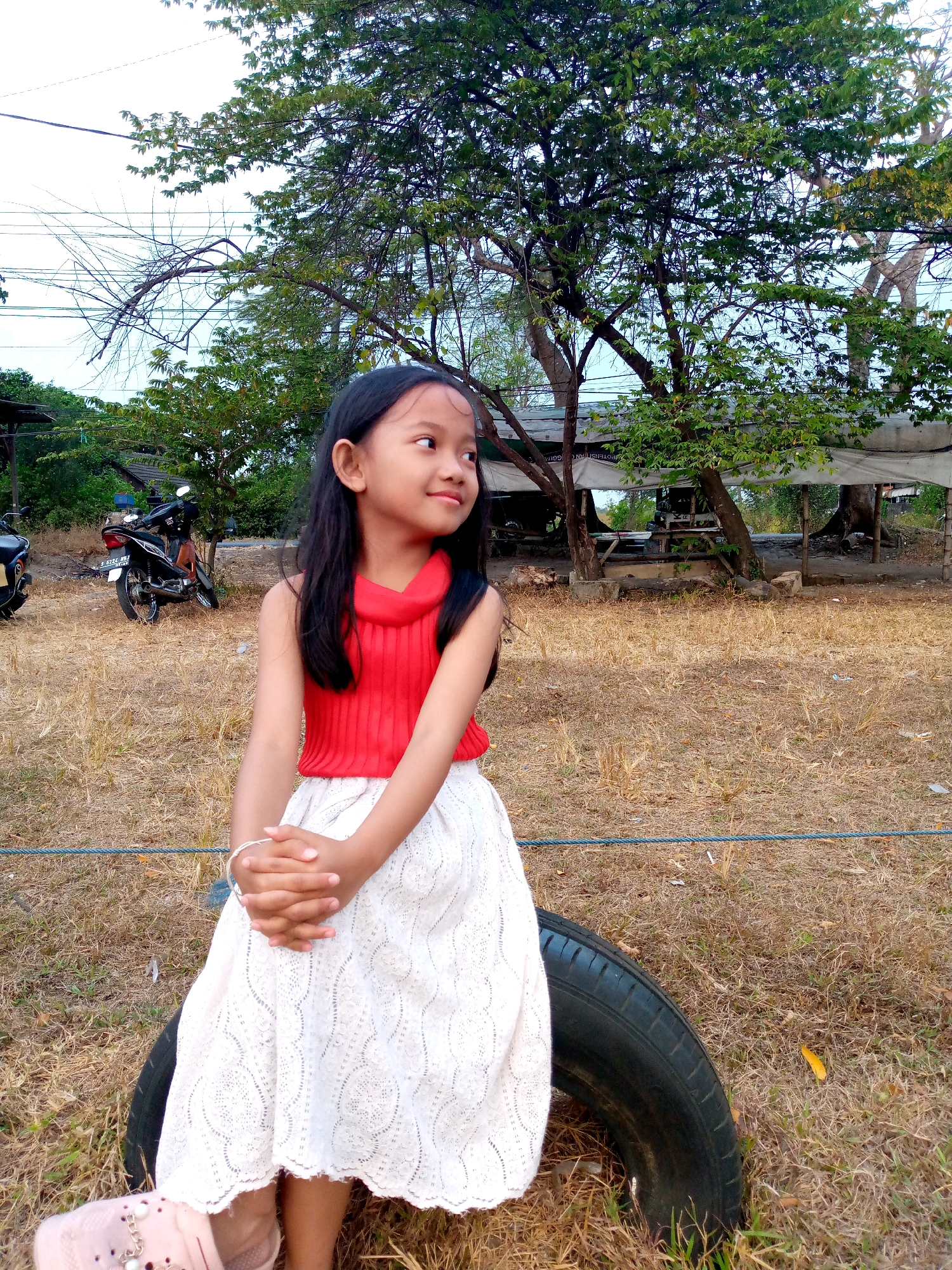 A young Indonesian girl with long, dark hair is sitting on a tire outdoors. She is wearing a red sleeveless top and a white lace skirt. Her hands are clasped in her lap, and she is looking to her right. The background shows dry grass, trees, and a motorcycle parked on the left. A wooden structure with a canopy is visible in the background. The scene appears to be in a rural or semi-rural area.