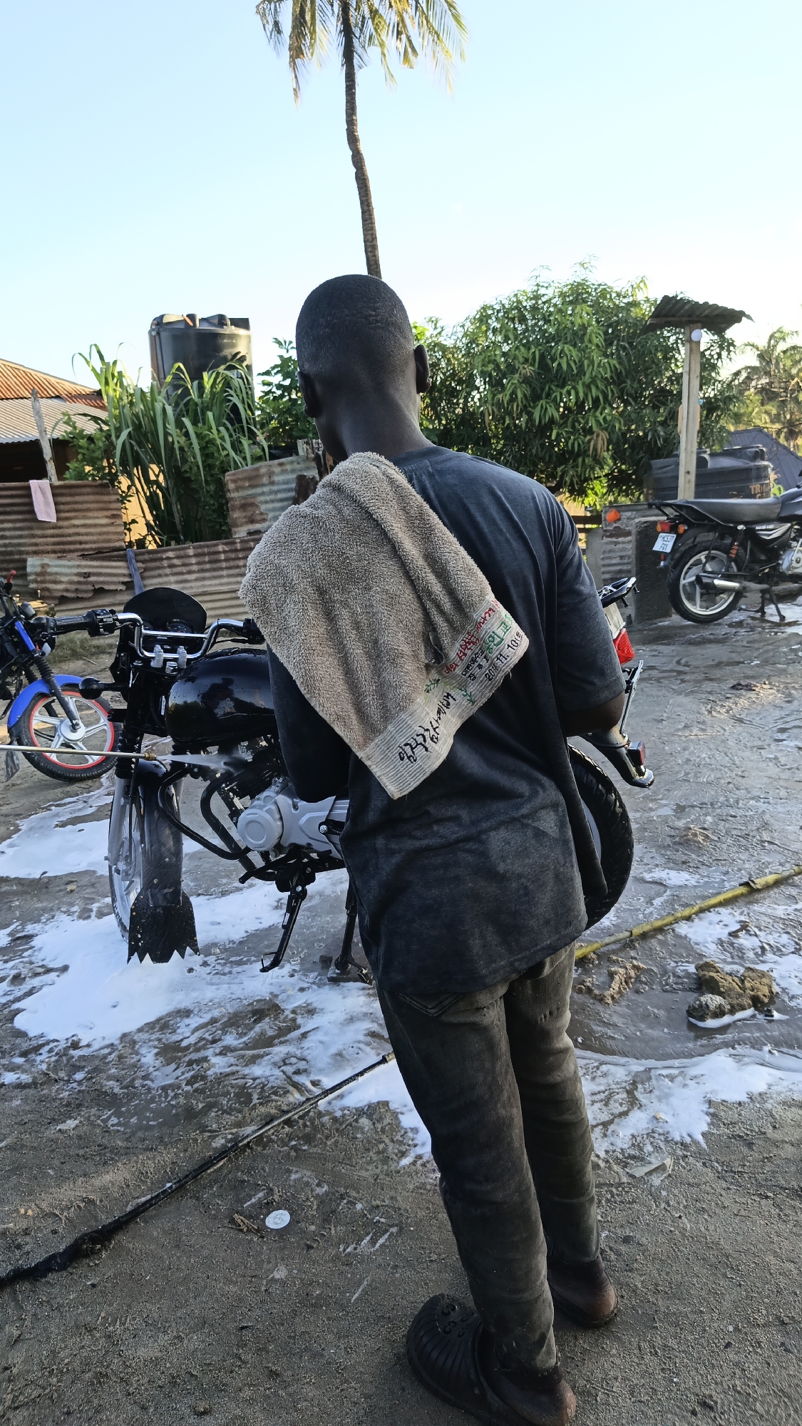 A man stands facing a motorcycle in what appears to be a residential or semi-rural area. The man is dark-skinned, with short hair, and is wearing dark clothing (a t-shirt and jeans) and black Crocs. He has a light-colored towel draped over his left shoulder.
The motorcycle in front of him is black and silver, standing on a kickstand. There are soapy suds on the ground around it, suggesting it is being washed. A second blue motorcycle is visible in the background to the left, and another black motorcycle to the right.
The environment includes buildings with corrugated metal roofing and a water tank in the background, along with trees and vegetation. A palm tree is visible in the center of the frame. The ground is a mix of sand and wet concrete or pavement. The lighting suggests it is daytime.
While I cannot confirm the location within Dar es Salaam specifically, the presence of corrugated iron buildings, motorcycles, and a palm tree are consistent with typical scenes in Tanzania. The scene depicts the mundane activity of washing a motorcycle.