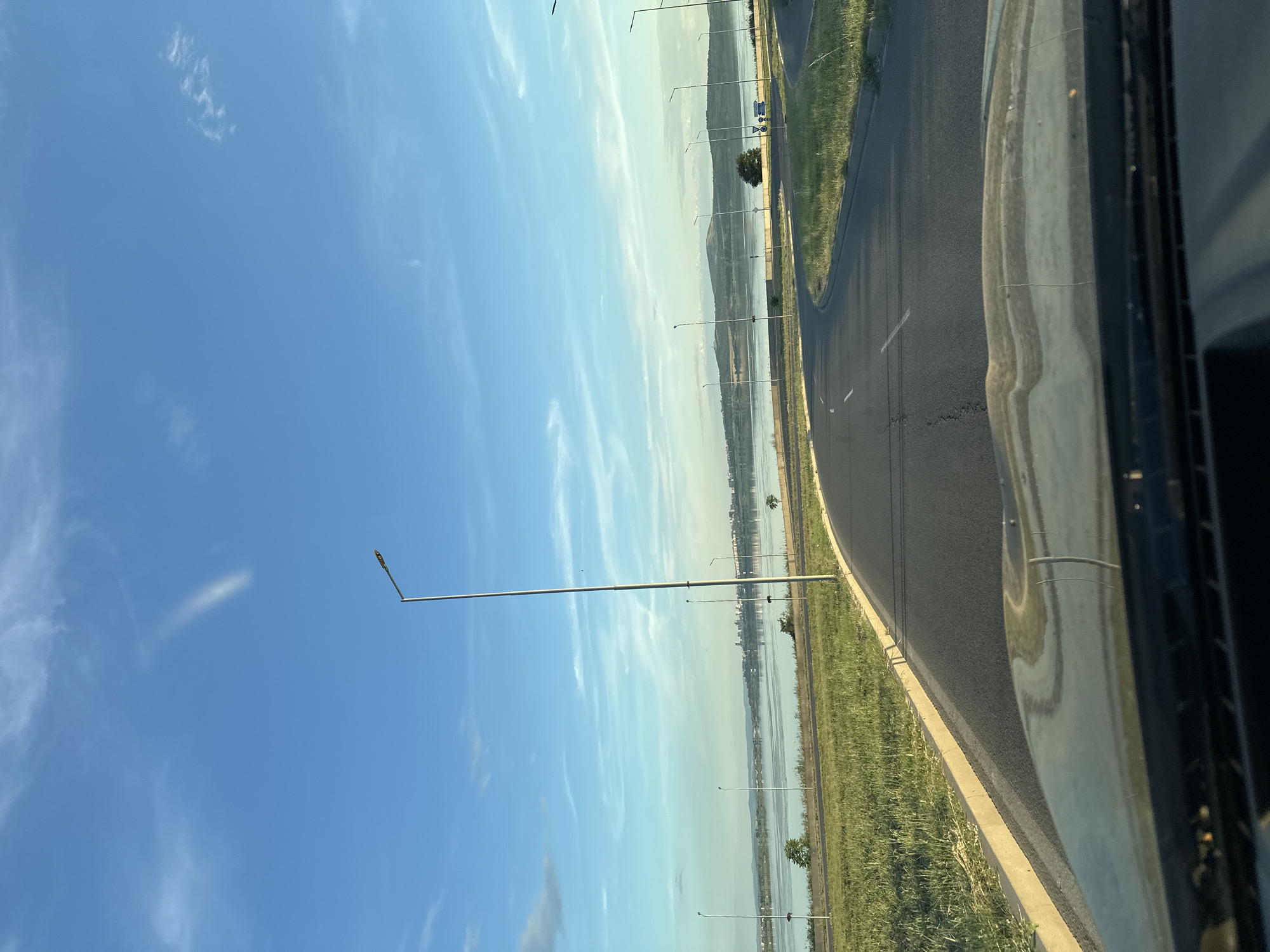 The image depicts a view from a car driving on a road with a body of water to the left, lined by grassy terrain and a sidewalk. The road curves to the right and continues towards a distant city skyline. Several lampposts are visible along the roadside, and the sky is a clear blue with scattered clouds, some wispy and others more defined. The overall impression is of a scenic drive along the coast or a large lake, with a suburban or semi-urban landscape.