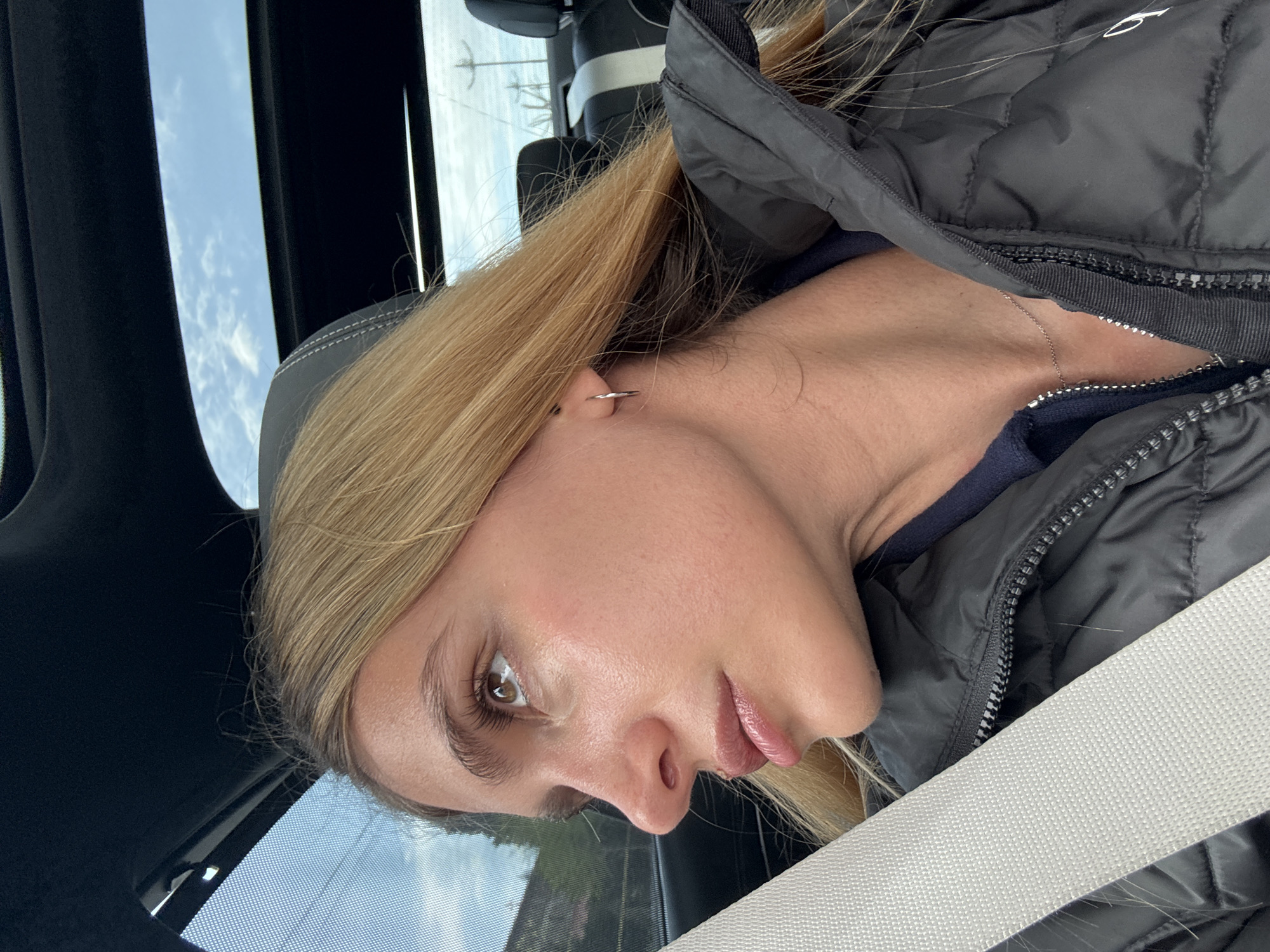 A woman with blonde hair is seated in a car, wearing a dark puffer jacket and a seatbelt. The car's sunroof provides a view of a cloudy sky. The location is Unknown, Unknown.