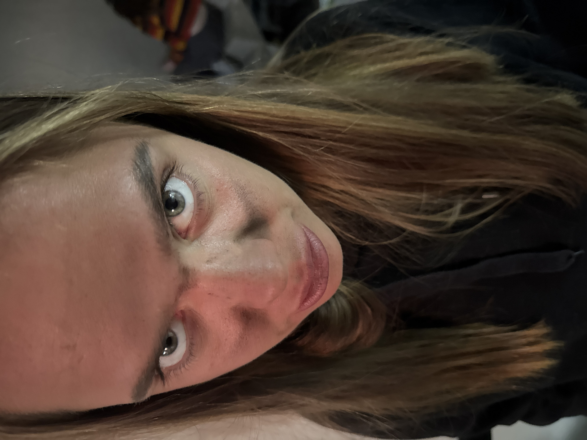 A close-up, tilted portrait of a person with light skin, light brown hair, and greenish-grey eyes. The person is looking directly at the camera with a slightly raised eyebrow expression. The person is wearing a dark-colored garment around their neck. The background is indistinct and out of focus. The image appears to have been taken indoors with artificial lighting. The location in Bol'shoye Kozino, Russia is not discernible from the image.