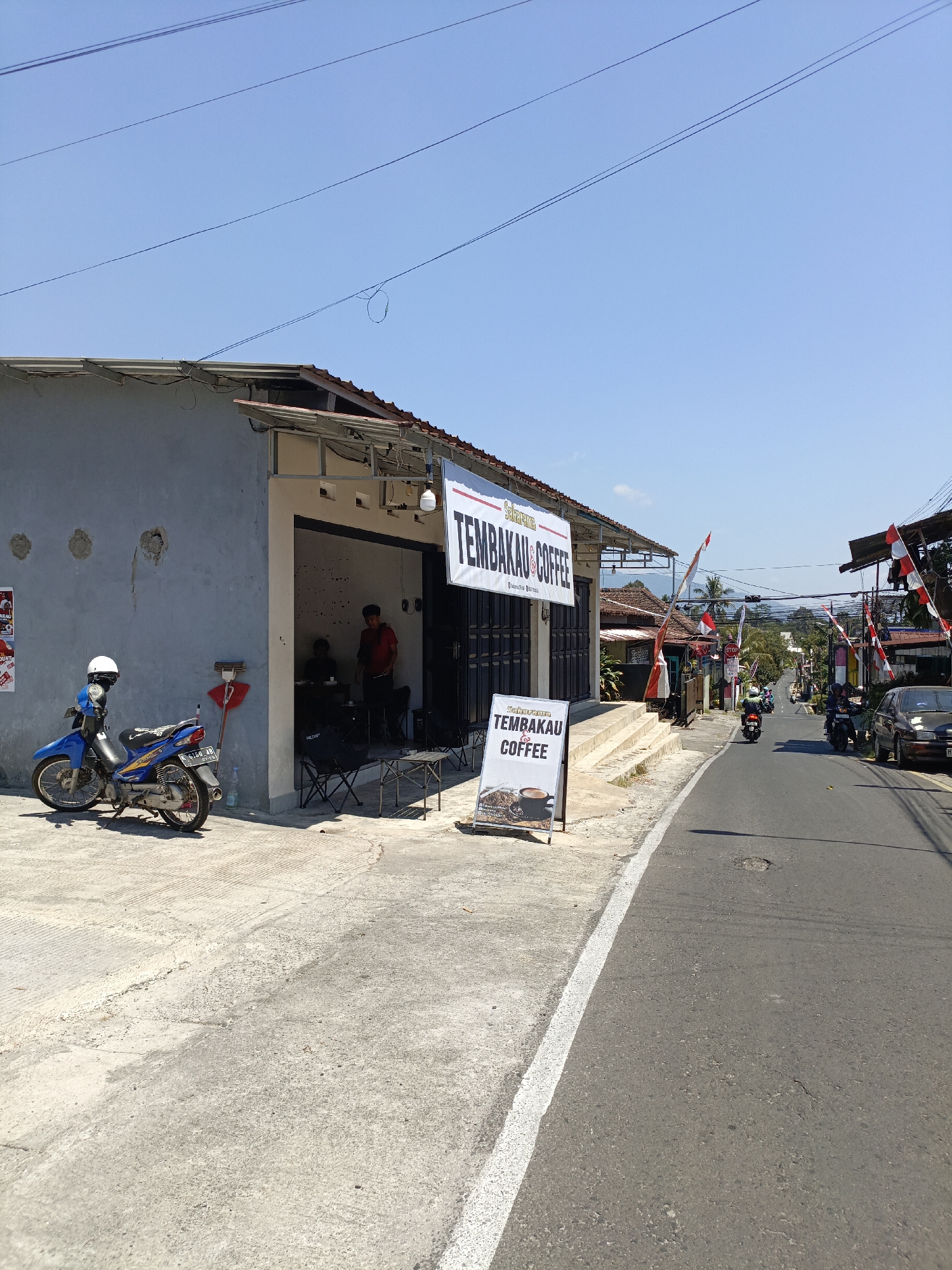 The image depicts a street scene in Salatiga, Indonesia, focusing on a coffee shop named "TEMBAKAU COFFEE." The coffee shop appears to be a small, open-fronted establishment with a gray exterior. A blue motorcycle with a white helmet parked in front of it is visible to the left of the entrance. Inside the shop, two individuals can be seen, one standing and wearing a red shirt, while another is seated. Two folding chairs and small tables are placed outside the entrance. A white A-frame sign with the coffee shop's name and an image of coffee is positioned on the sidewalk. The street is paved and has a white lane marking. Several Indonesian flags are displayed along the street, suggesting a local event or celebration. Other buildings and vegetation line the street, and in the distance, hills and a clear blue sky are visible. The overall scene suggests a sunny day in a residential or commercial area.