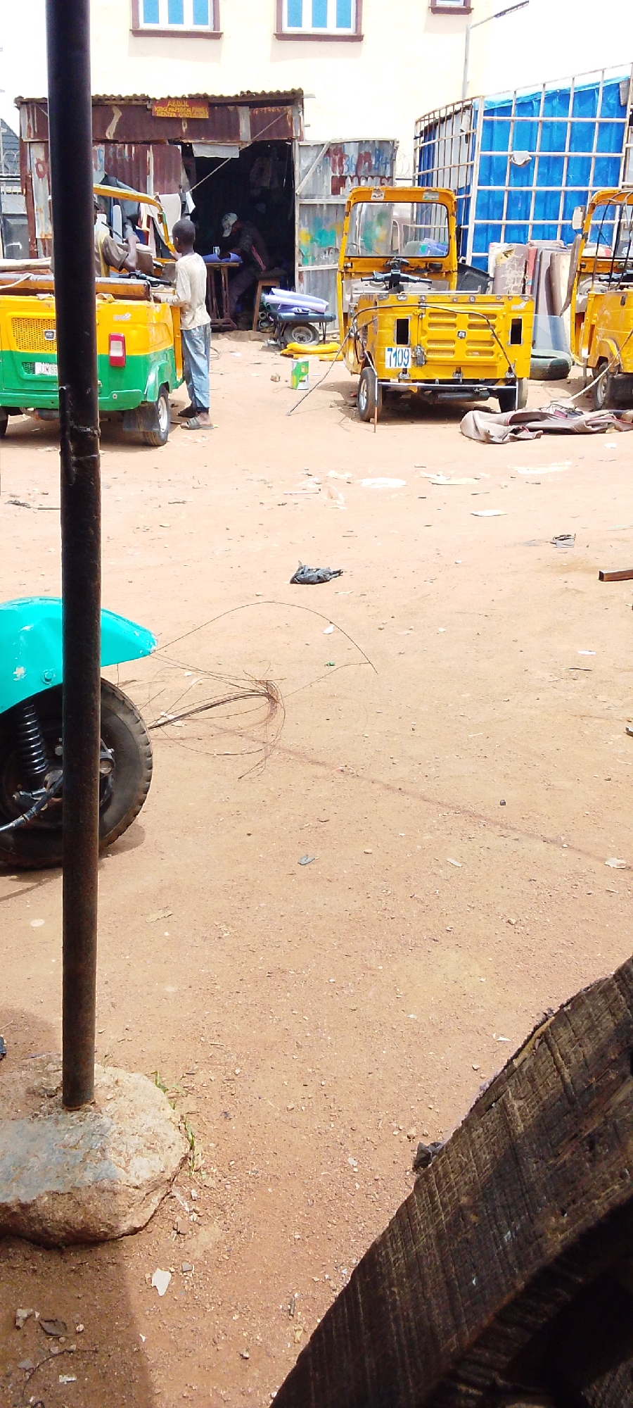 This photo was taken during daylight in Jos, Nigeria. The ground is made of sand. The scene is cluttered with auto rickshaws. One person stands in front of a building, near auto rickshaws that are yellow and green. Another person works inside of the building. The building looks like a paper factory. A pole stands in the foreground with a light blue auto rickshaw to the left.