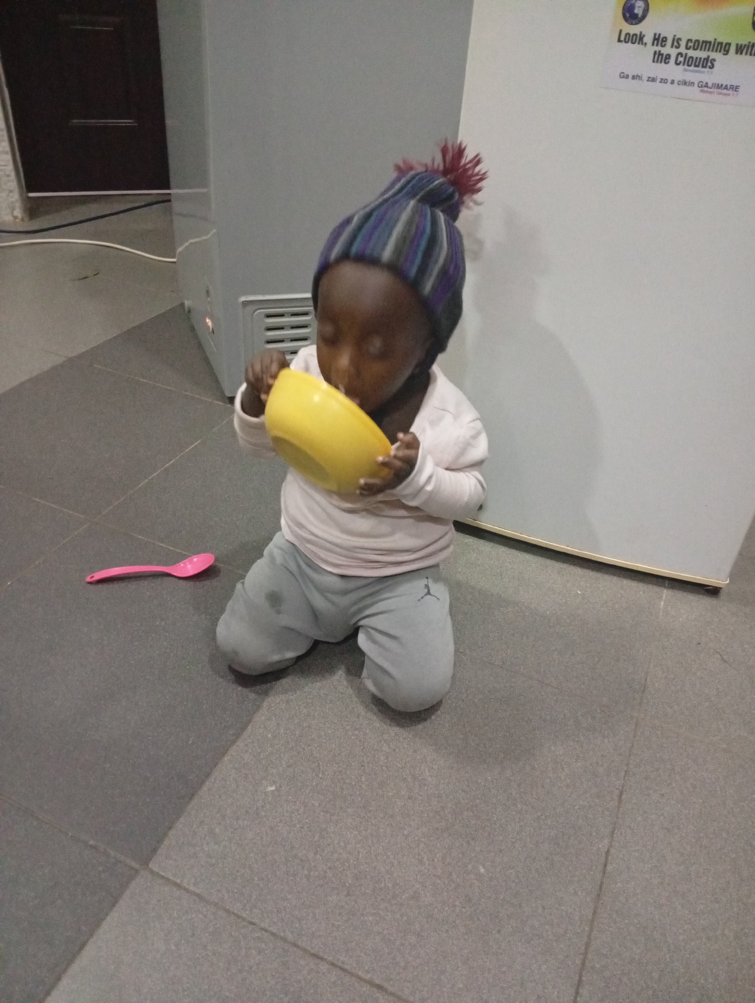 A young child wearing a striped beanie with a pom-pom is kneeling on a tiled floor, drinking from a yellow bowl. The child is also wearing a light-colored long-sleeved shirt and grey pants. A pink spoon is lying on the floor next to the child. In the background, a light-colored appliance, possibly a refrigerator, is visible. A dark door and a white cable are also present in the background. A poster with text that reads "Look, He is coming with the Clouds" and other inscriptions in a foreign language is affixed to the appliance. The scene appears to be indoors, possibly in a home or community center in Gura Top, Nigeria.
