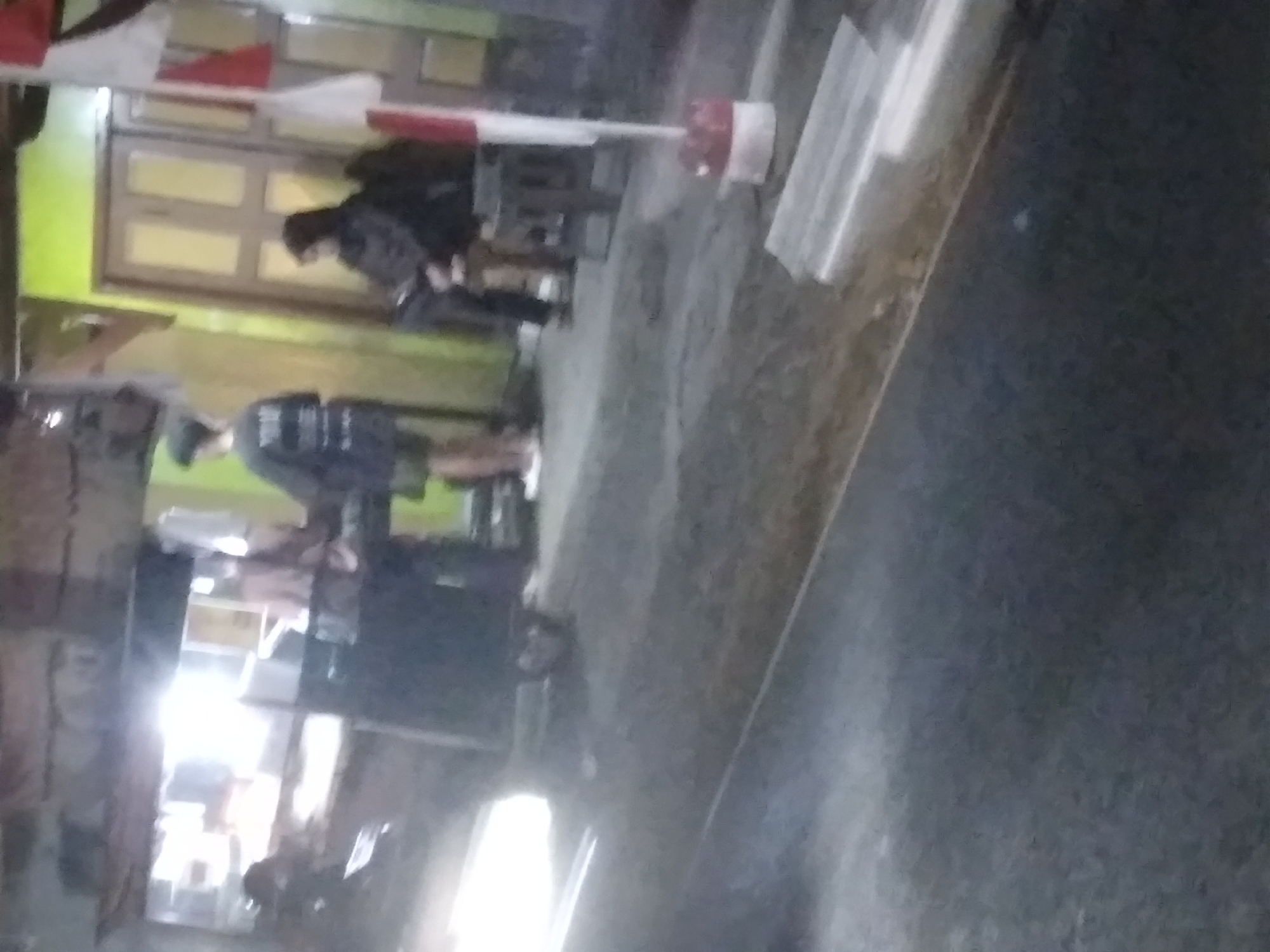 The image depicts a street scene at night in Sokaraja, Indonesia, likely near a food stall or small establishment. In the foreground, a blurry dark asphalt road is visible, with some white objects that could be street markings or debris. In the mid-ground, a brightly lit food stall or counter is present, with a person standing behind it, possibly preparing food or serving customers. To the right of the stall, a person is seated on a stool, appearing to be a customer. Further to the right, a wooden door painted yellow with white panes is set into a lime green wall. A red and white flag on a pole is also visible near the door. In the background, indistinct shapes suggest buildings or structures. The lighting is harsh and reflective, creating a somewhat underexposed and grainy appearance. The overall atmosphere is casual and appears to be a typical evening scene in a commercial area of the city.