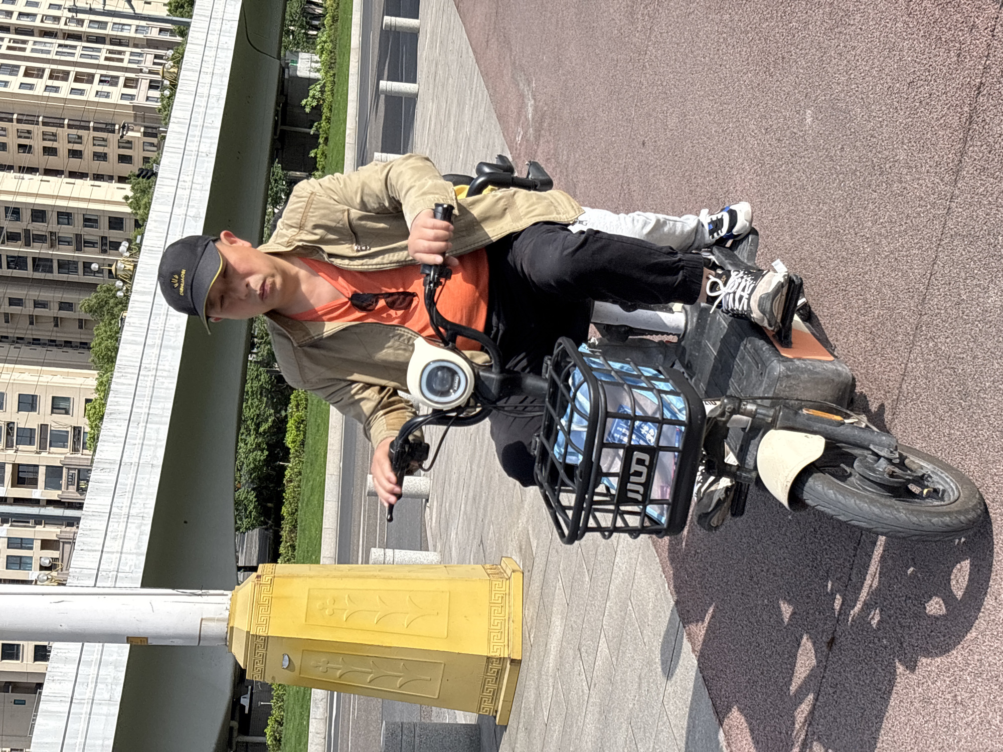 A man in a tan jacket and black pants rides an electric scooter on a sidewalk. The man wears a black baseball cap and an orange shirt, and sunglasses rest on his chest. A child rides with him. A black wire basket sits on the front of the scooter. In the background are modern buildings, trees, a bridge, and an ornate yellow post. The location is identified as Jiangjin, China.