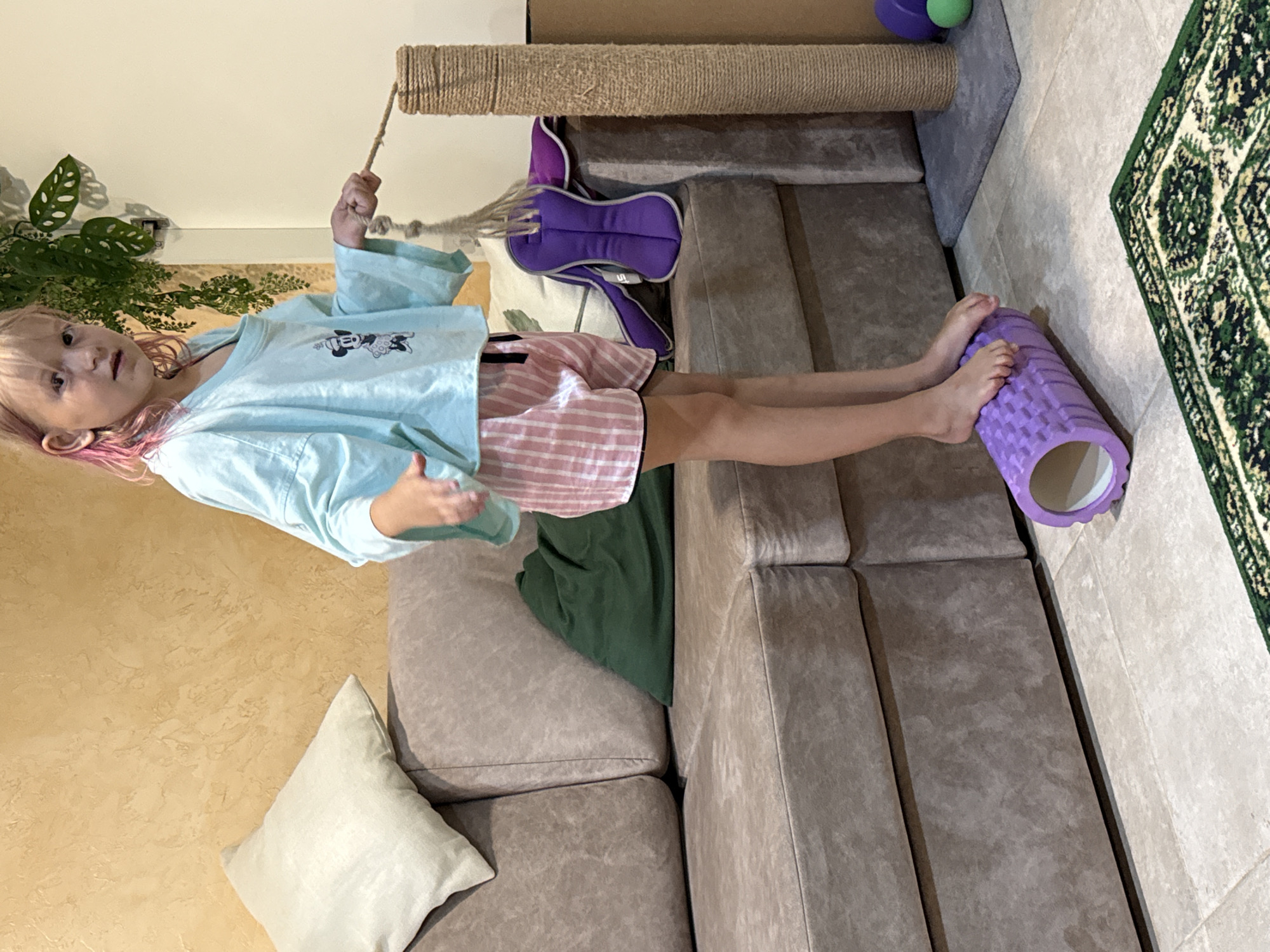 A young girl with blonde hair with pink streaks stands on a purple foam roller in what appears to be a living room. She is wearing a light blue oversized shirt with a Mickey Mouse graphic and pink and white striped shorts. Her feet are positioned on top of the roller, which is placed on a tiled floor. To her left is a large, taupe-colored sofa with several pillows, including a light beige one and a dark green one. To her right, a tall, brown sisal cat scratching post stands next to a purple stuffed toy and a green ball. In the background, a textured yellow wall is visible, and a leafy green plant hangs from a hook. A portion of a green and white patterned rug is visible in the foreground. The girl is holding onto a rope that is attached to the cat scratching post.