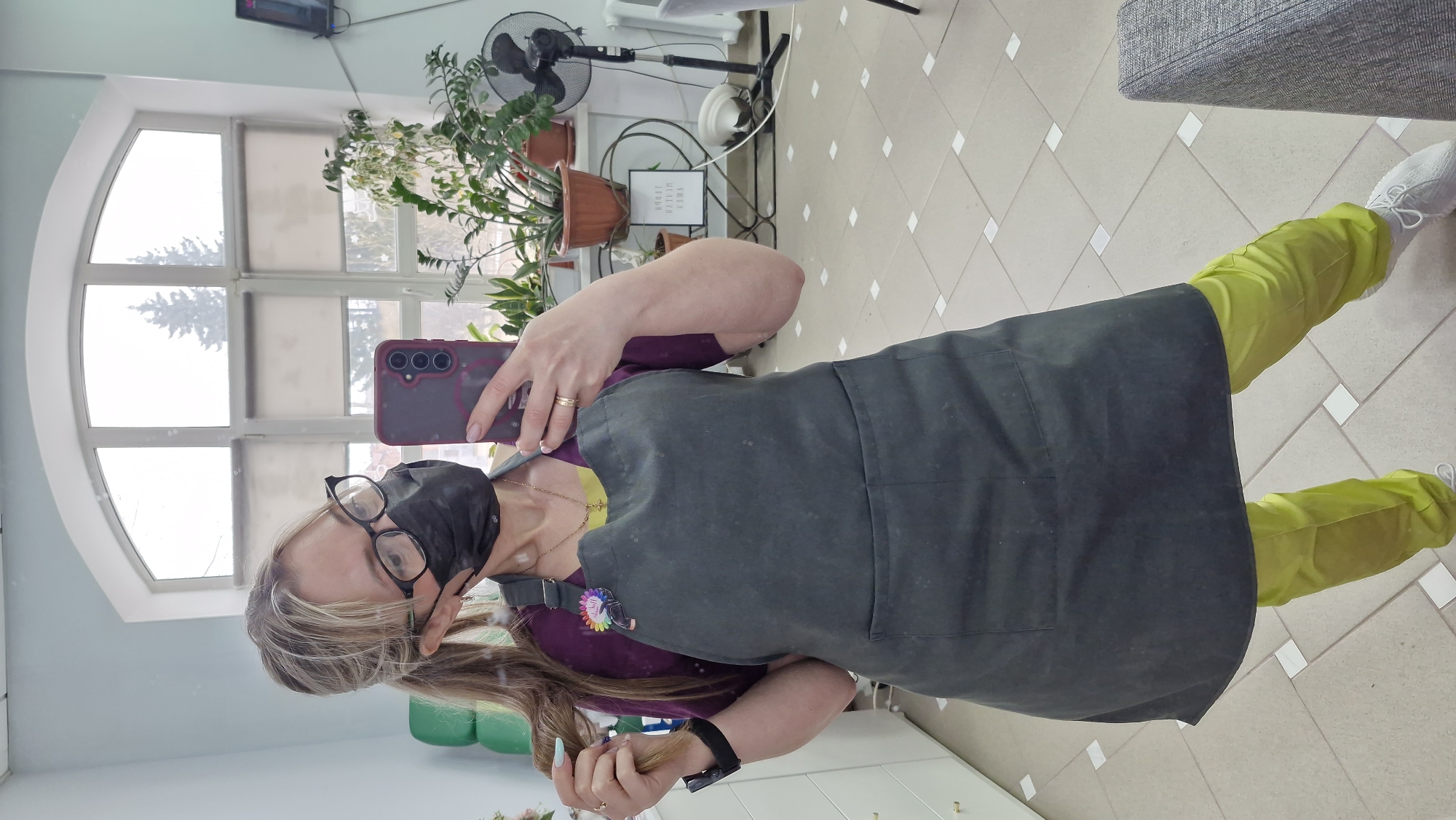 A woman wearing eyeglasses and a black face mask is taking a selfie in what appears to be a salon setting. She is wearing a dark gray apron over a purple shirt and light green pants. She holds a maroon-colored smartphone. The background features a large, arched window, plants in pots on a shelf, and a black standing fan. The floor is tiled. The location appears to be in an interior space in Otchor, Russia based on information available in the image.