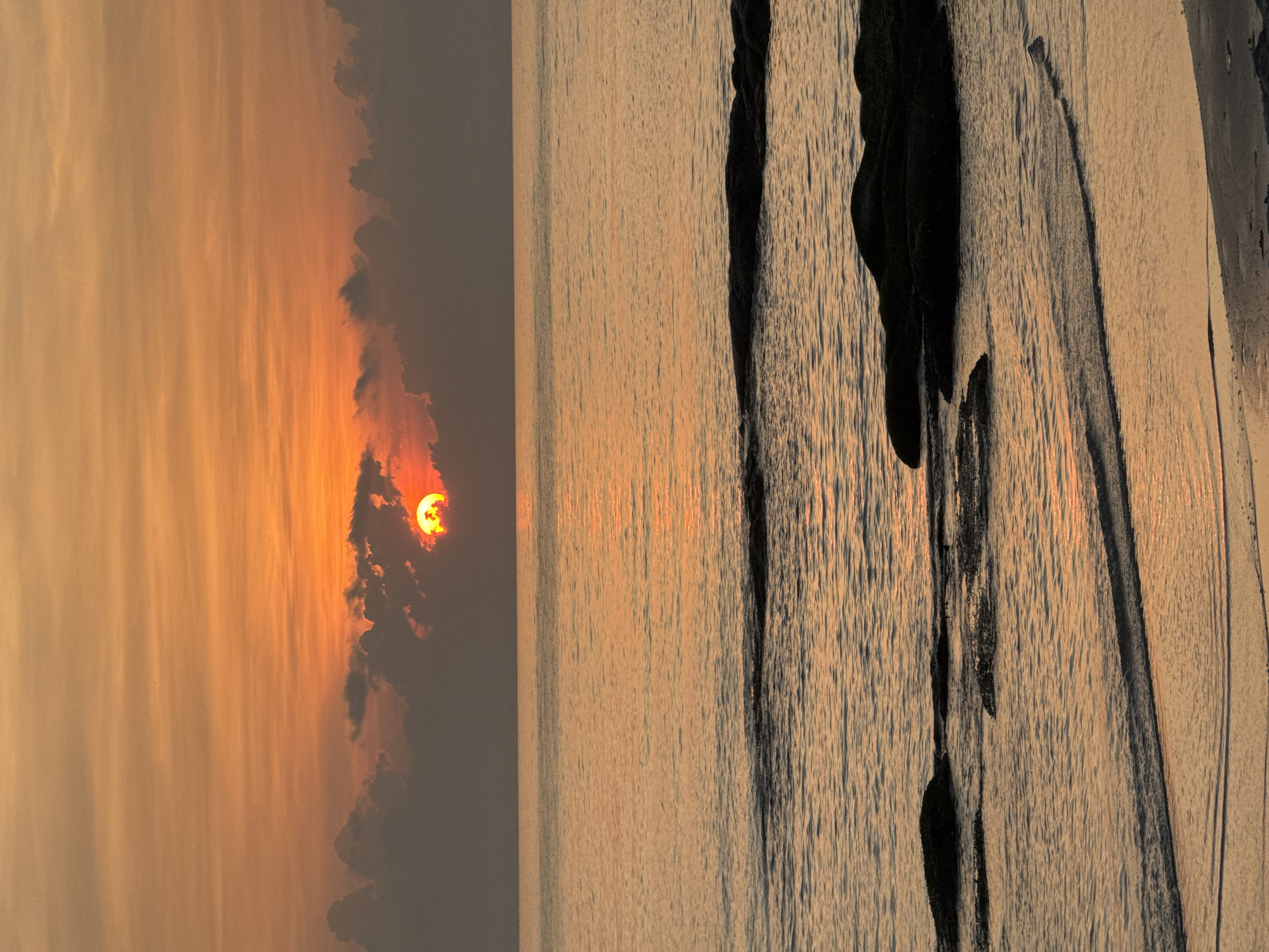The image shows a view of Chaweng Beach in Baan Chaweng, Thailand, at sunset. The sun, appearing reddish-orange, is positioned on the left side of the frame, partially obscured by clouds. The sky around the sun is filled with orange and yellow hues from the setting sun. To the right of the sunset is the ocean, reflecting the same golden colors. The texture of the water is visible due to slight ripples. Dark, irregular shapes are visible on the right side of the frame, possibly rocks or natural formations along the beach.
