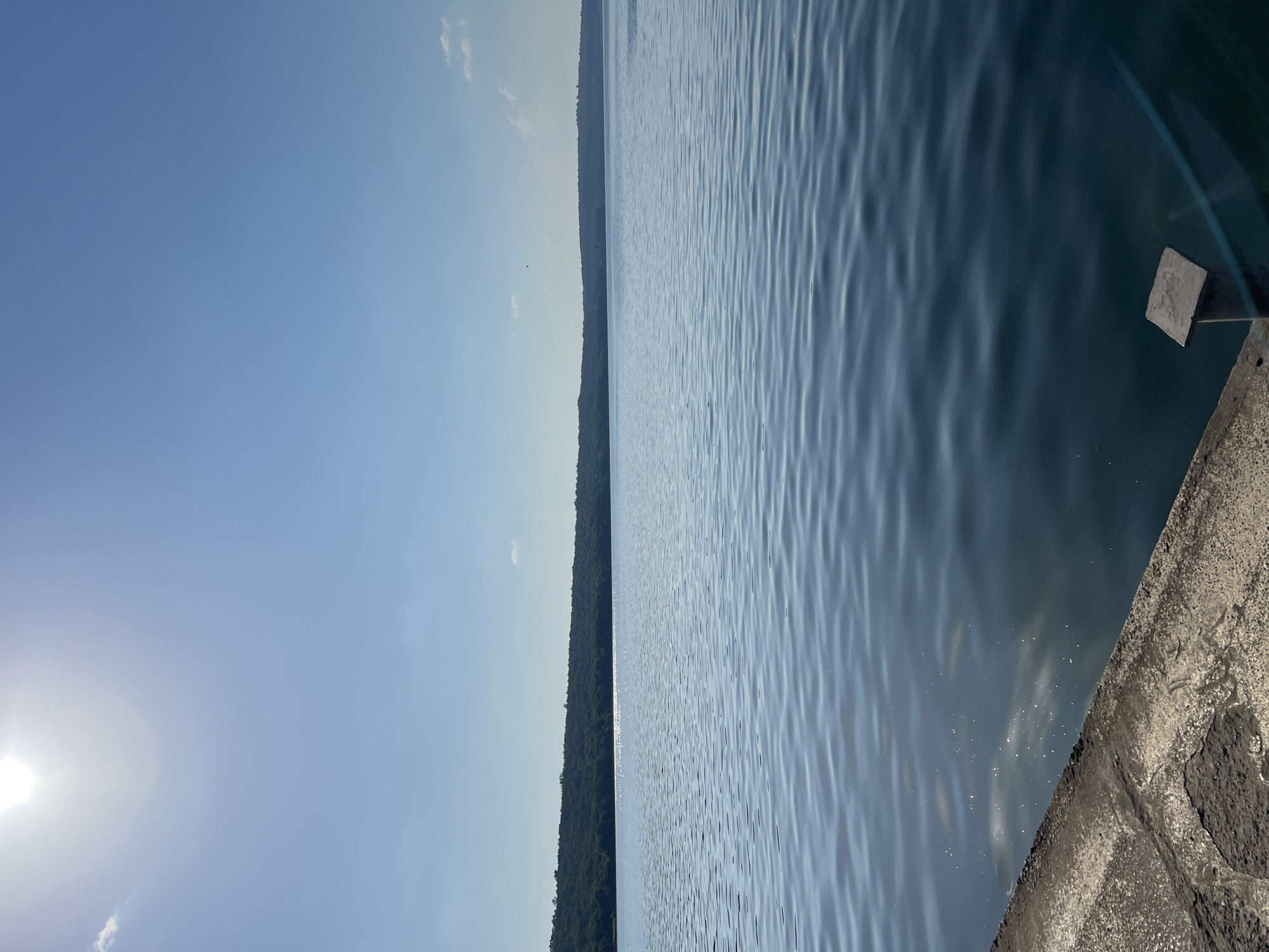 The image shows a body of water with ripples, likely a lake or river, under a clear blue sky. The sun is visible in the upper left corner, suggesting it is daytime. On the right side of the image, there is a concrete embankment or wall with some texture. In the distance, a treeline or forested area is visible along the horizon, separating the water from the sky. The overall scene is serene and appears to be an outdoor landscape. There is no visible text in the image. The orientation of the image is sideways, with the horizon running vertically.