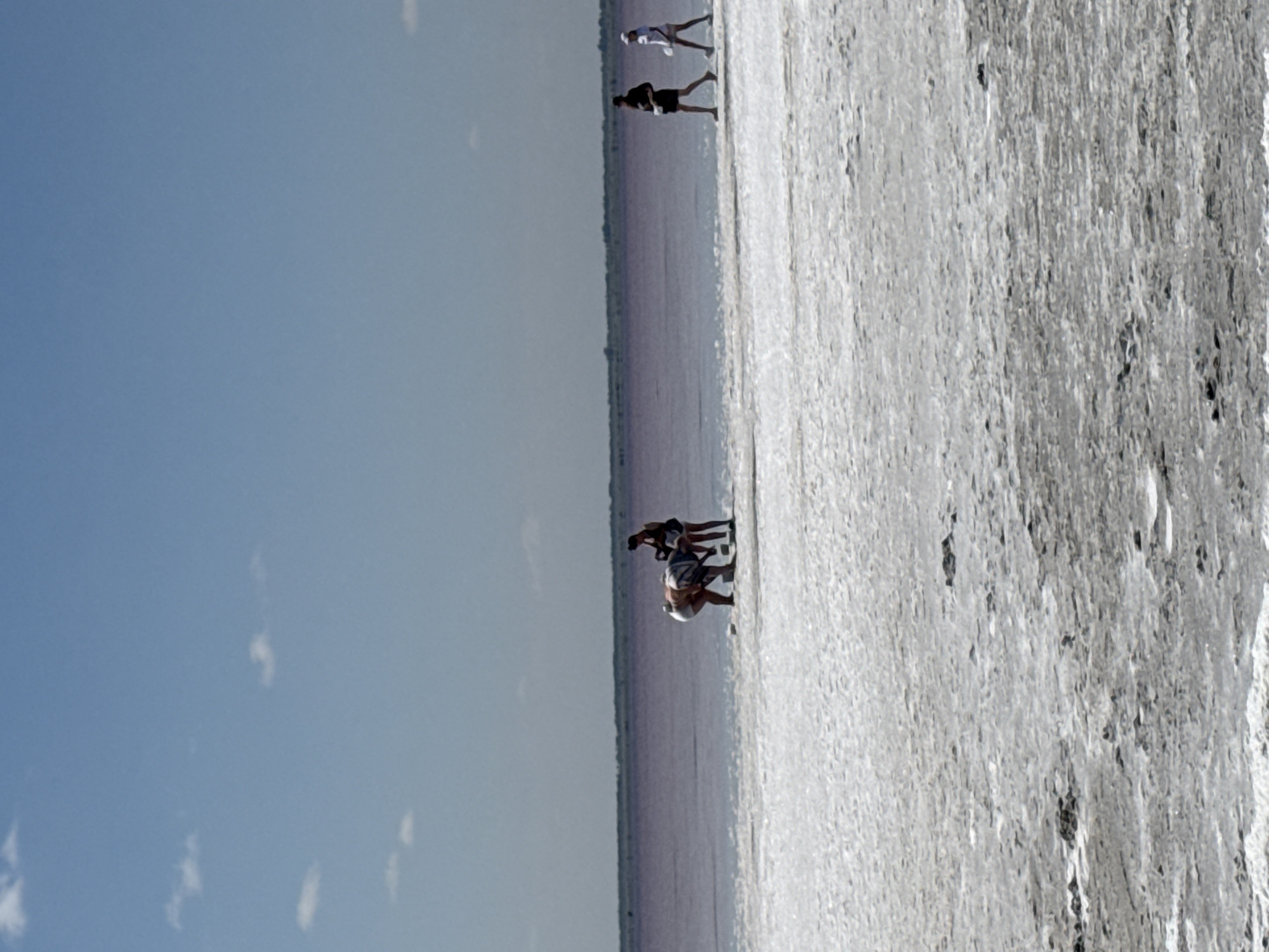A wide shot captures a sunny day at a salt lake in Бурсоль, Russia. The sky is a clear, pale blue with sparse white clouds. The foreground is a white, textured salt flat. Beyond the salt flat lies a calm, pale pink body of water, characteristic of the salt lakes in the region. In the distance, a dark, thin line of vegetation marks the horizon.

In the middle ground, several people are visible near the edge of the salt flat and the water. On the left, two individuals are bent over, appearing to be interacting with the salt or water. One person is wearing a striped top and shorts, while the other is in a white top and shorts. To the right of them, a person wearing a dark top and shorts walks away from the camera. Further to the right, another person in a white shirt and shorts walks toward the left, also facing away from the camera. The people are walking or interacting individually, with no apparent direct interaction between the groups. The overall scene depicts a tranquil day at the unique natural environment of the salt lake.