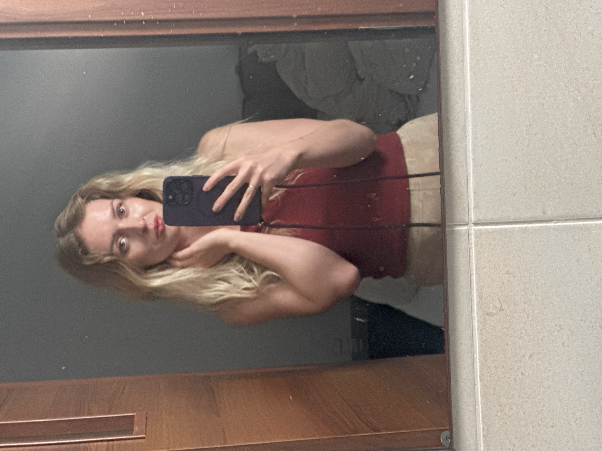 A fair-skinned female with long blonde hair is taking a mirror selfie in a bathroom. She wears a burgundy tank top and light-colored pants. She is holding a dark-colored smartphone up to her face with her right hand. She has a ring on her ring finger. The bathroom has a wooden-framed mirror, light-colored tile walls, and a gray wall reflected in the mirror. The location is unknown and there is no information to indicate that the image was taken in Petah Tikva, Israel.