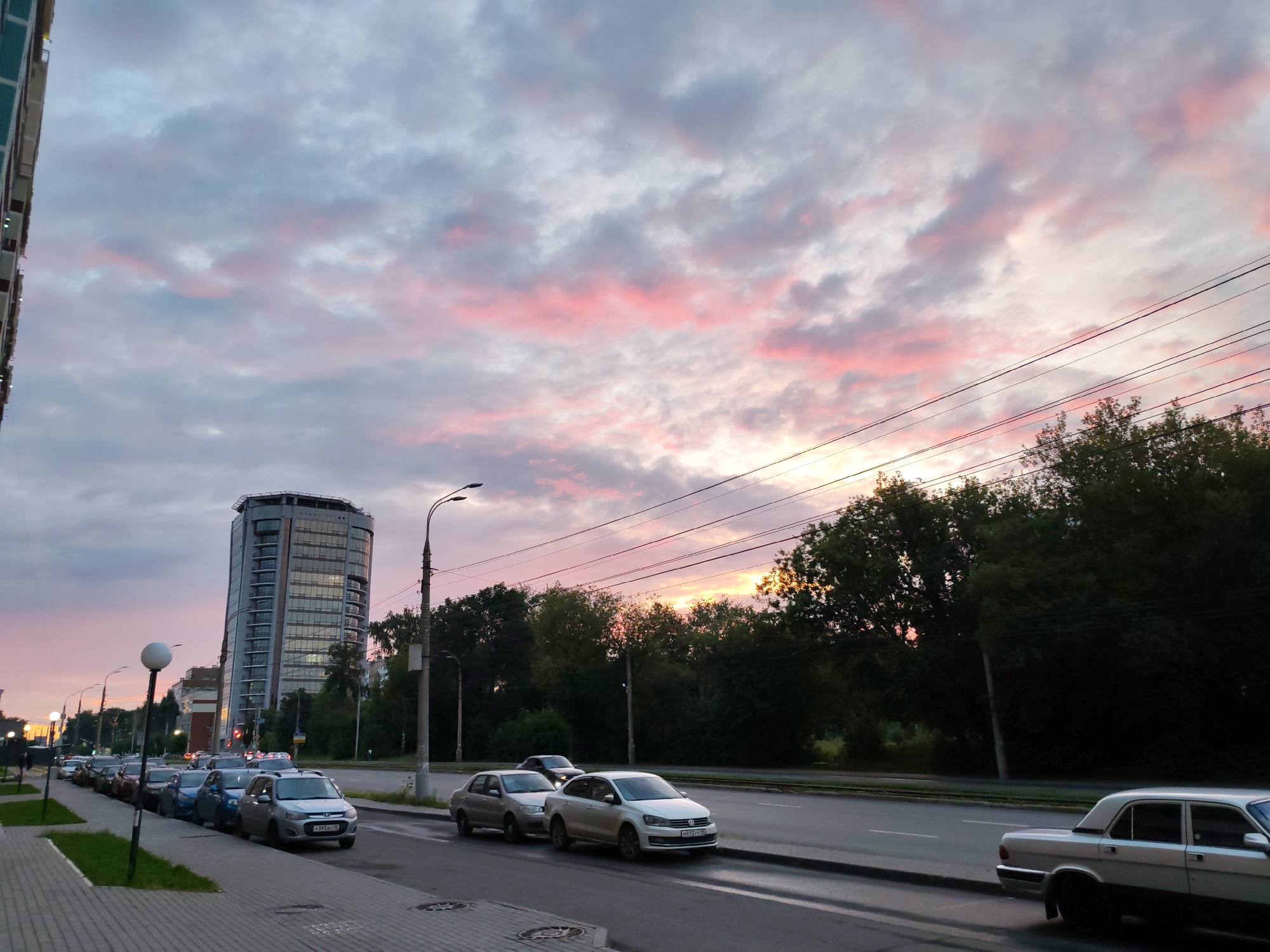 The image depicts a street scene in Izhevsk, Russia, during what appears to be sunset or dusk. A multi-story, modern building with a curved facade dominates the skyline on the left side of the image, reflecting the warm hues of the sky. A row of cars is parked along the right side of a wide paved road. The sky is filled with dramatic clouds, tinged with pink and orange, suggesting the setting sun. Dense green trees line the opposite side of the road, creating a visual barrier between the street and the background. Power lines are strung across the upper portion of the image, intersecting with the sky. The foreground shows a sidewalk and street lamps. There are no people visible in the image.