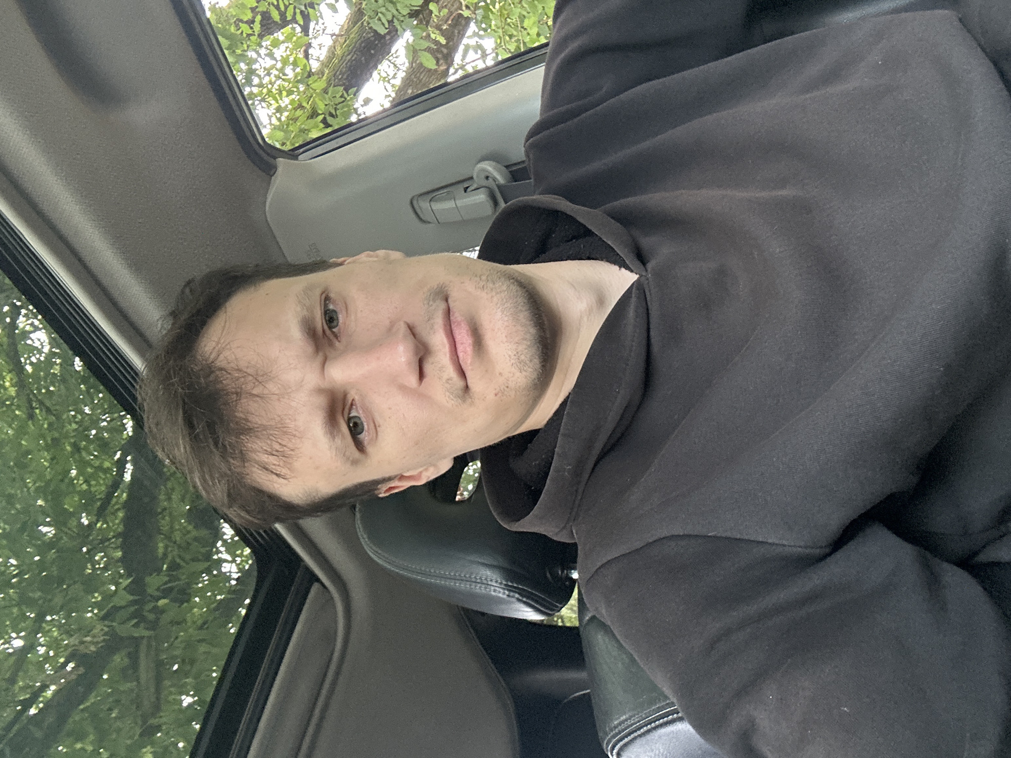 A selfie of a man is taken from inside a car, looking directly at the camera. He is wearing a black hooded sweatshirt. The car's sunroof is open, revealing green foliage and tree branches from above. The interior of the car is visible, including a black leather headrest. The background outside the car is blurry but suggests an outdoor, possibly wooded, environment. The image appears to be taken during daylight. The location is identified as Makiv, Ukraine.