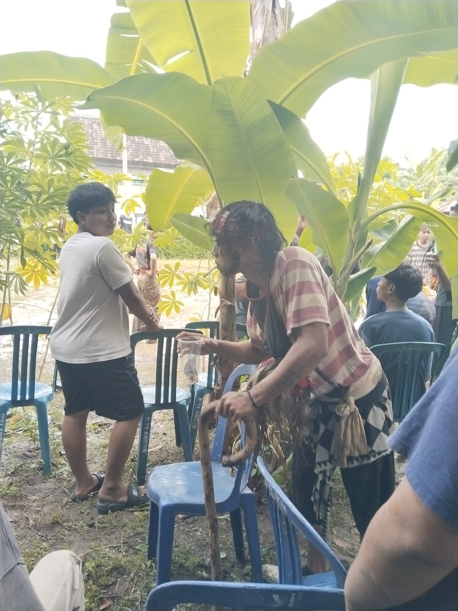 The image, taken outdoors in Karang Tengah, Indonesia, shows a gathering of people around a banana tree. One man is in a striped shirt and dark pants with a checkered scarf-like accessory tied at the waist, holding a walking stick. He has tattoos on his left arm and his hair is disheveled. He appears to be holding a clear plastic cup. Beside him, a younger man in a white t-shirt and dark shorts stands with one hand on a light blue plastic chair. Other people are visible in the background, some seated on similar chairs. The ground appears damp, and there are other trees and foliage in the background, along with a glimpse of a building in the distance. The atmosphere is relaxed and casual.