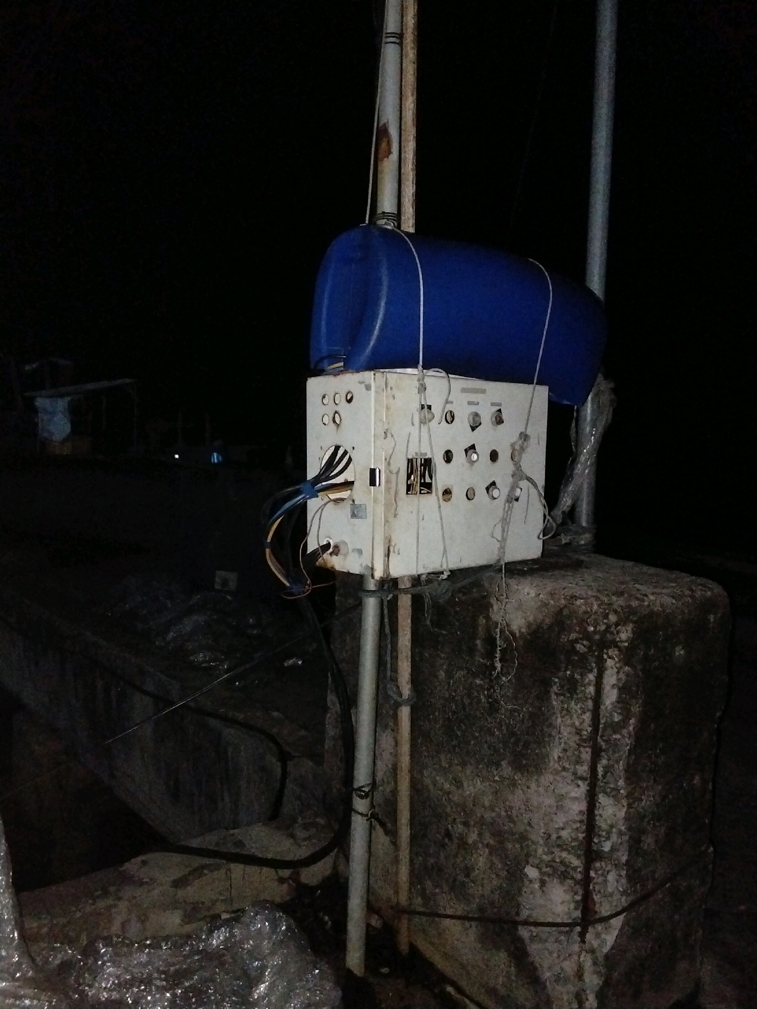 The image shows an outdoor scene at night, likely in Moro, Indonesia. A weathered white electrical control box is mounted on a metal pole, which is secured to a large, moss-covered concrete block. A blue plastic barrel is strapped to the top of the control box. Wires are seen entering and exiting the box, with some loose and exposed. The background is dark and out of focus, with hints of structures and possibly water in the distance, suggesting a harbor or coastal area. The overall impression is one of a functional, but unkempt, piece of outdoor electrical infrastructure.