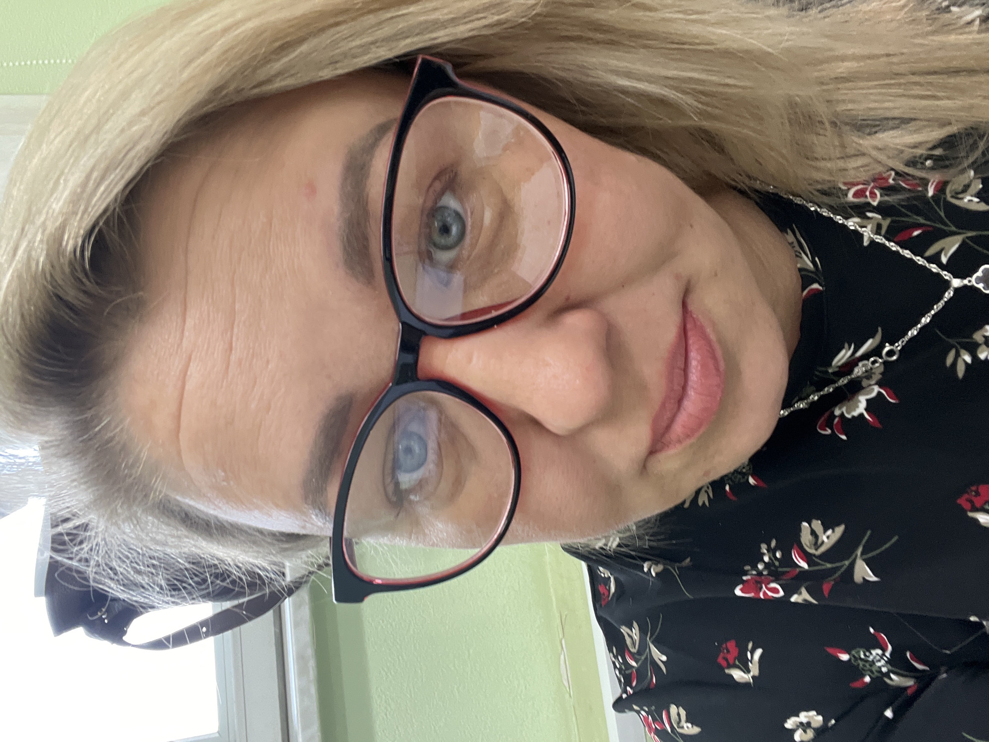 A woman with blonde hair and glasses is pictured in a close-up, slightly angled shot. She is wearing a black floral-patterned dress and a silver necklace. The background appears to be a light green wall. No specific elements indicate the location is Chaikovsky, Russia.