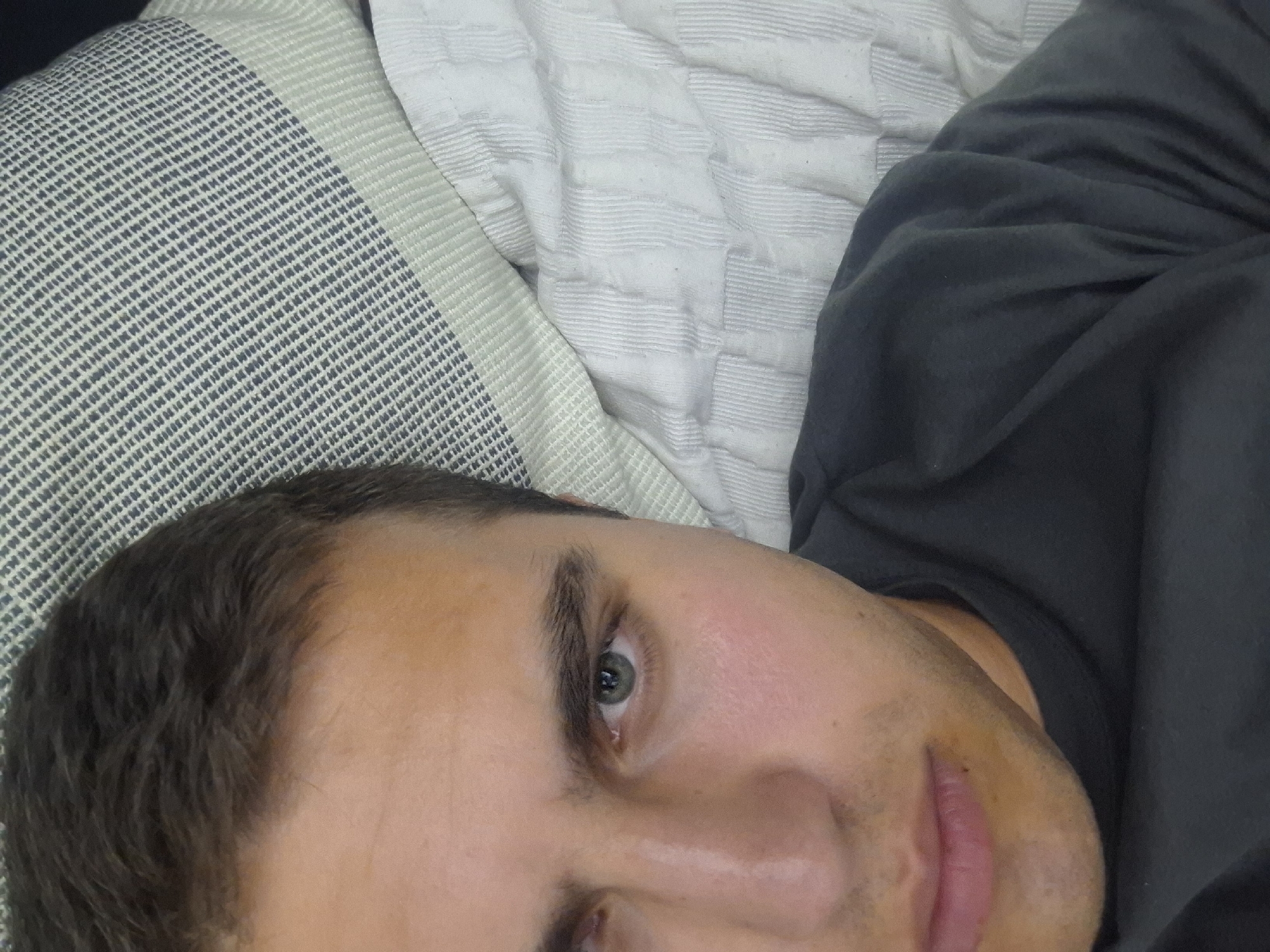 A close-up, high-angle, selfie-style shot shows the left side of a fair-skinned individual's face and upper torso. The person has dark brown hair, light brown eyebrows, and green eyes. The right side of the face is partially obscured by a pillow with a patterned gray and white fabric. The individual is wearing a dark gray crewneck t-shirt. The background consists of a white, quilted surface, possibly a bed or sofa. The lighting appears to be artificial and is coming from the front and slightly above. The image was taken in Pleven, Bulgaria.