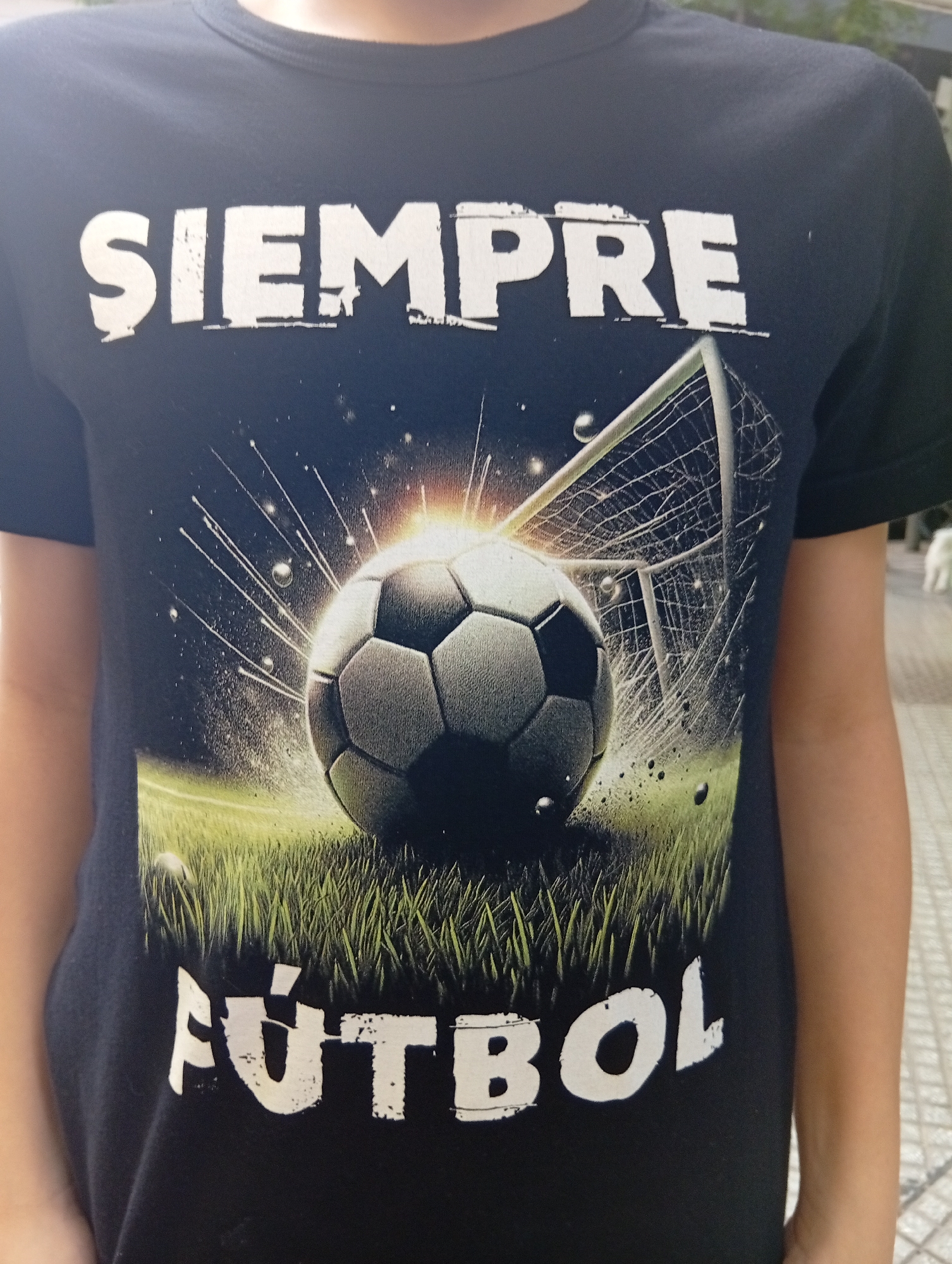 A person, likely a child or young adult, is depicted from the chest up, wearing a black short-sleeved t-shirt. The t-shirt features a dynamic graphic design centered on a soccer theme. A prominent soccer ball rests on vibrant green grass, radiating a bright, glowing light that emanates from its upper right side. This light illuminates the ball and its immediate surroundings, with sharp, energetic streaks extending outwards against a dark, speckled background, suggesting an impact or powerful motion. Partially visible behind the soccer ball and to its right is a white soccer goal with its net.
In large, white, distressed capital letters above the graphic, the word "SIEMPRE" is printed. Below the graphic, in a similar style, is the word "FÚTBOL," with an accent mark over the 'Ú'. The full text reads "SIEMPRE FÚTBOL," meaning "ALWAYS SOCCER" in Spanish.
The person's bare arms are visible on either side of the t-shirt. In the soft-focus background, a small, light-colored dog is partially visible on the far right. The ground in the background is paved with light-colored tiles or stones, suggesting an outdoor, possibly urban or residential, setting consistent with Buenos Aires, Argentina. The image captures a casual moment, highlighting the wearer's passion for soccer through the t-shirt's design.