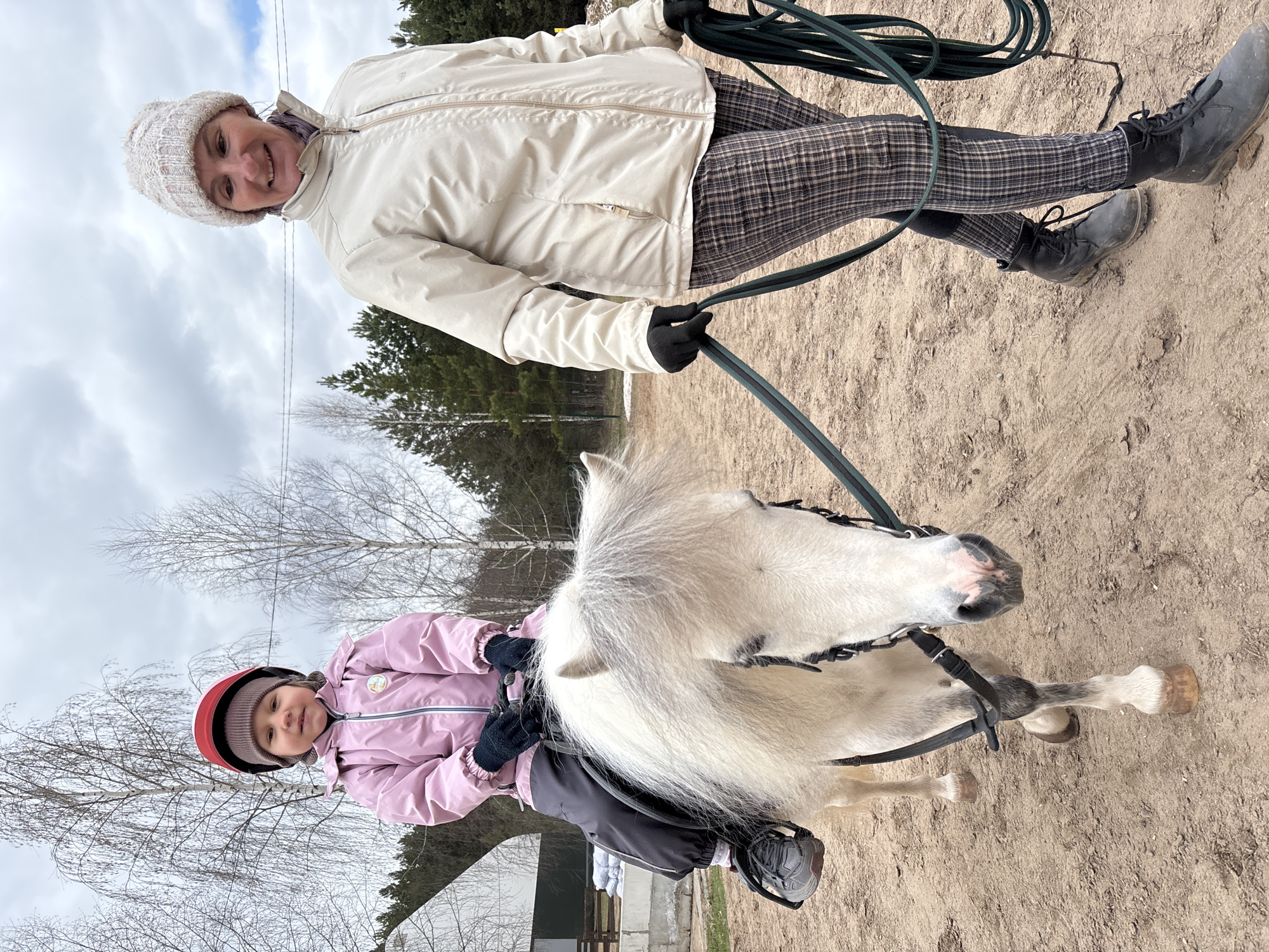 The image shows a woman standing on a sandy surface while holding a lead rope attached to a white pony being ridden by a child wearing a pink jacket and a red helmet. The woman is wearing a white jacket, plaid pants, black boots, a knit hat, and black gloves. The background includes trees and a cloudy sky. The image was taken in Русский Вожой, Russia.