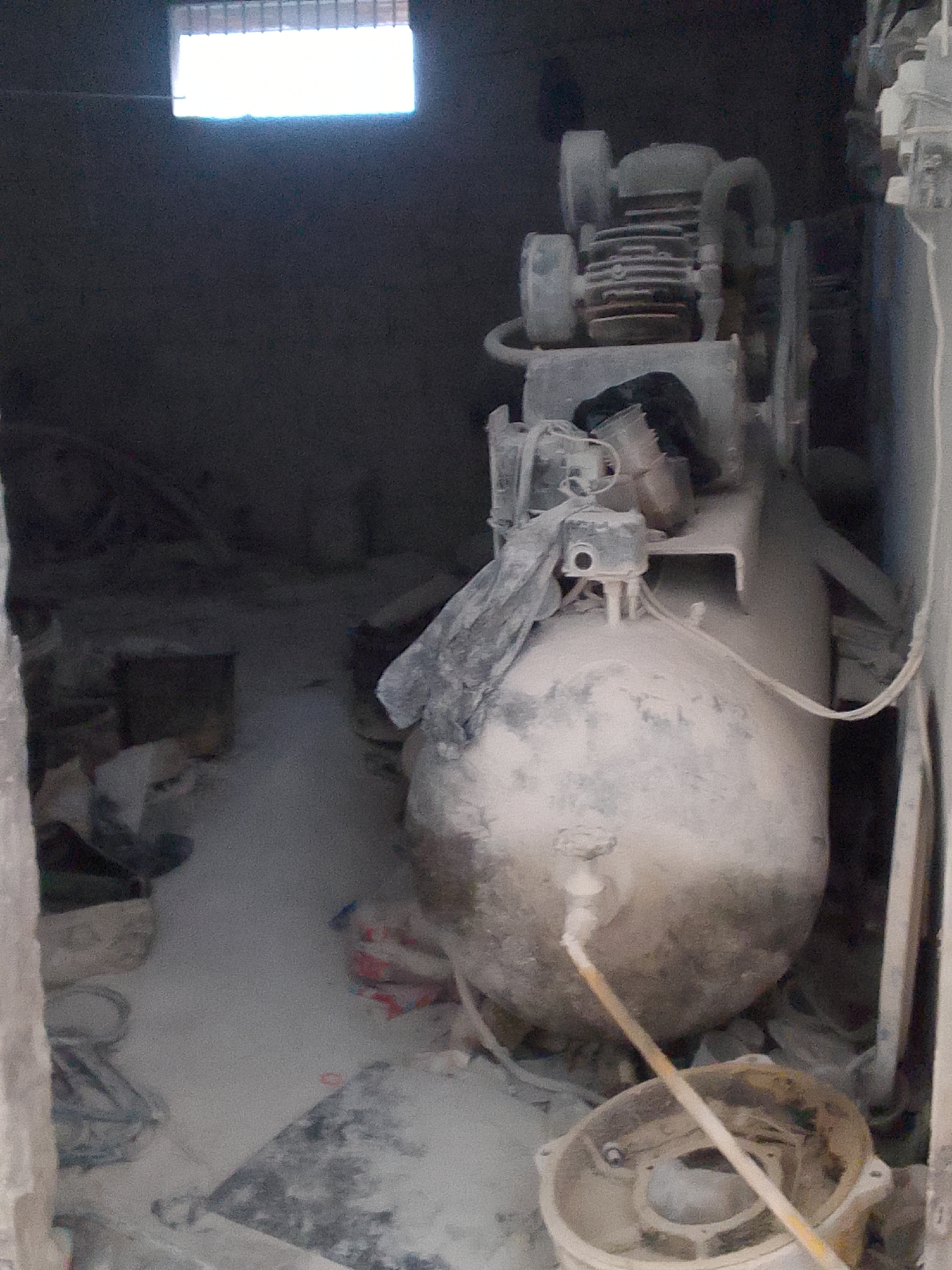 The image shows an interior scene, likely a workshop or storage area, covered in a fine white dust, suggesting recent work with powdered materials. In the foreground, a large, round, dusty tank dominates the right side of the frame, with a yellow tube running from its side down towards the bottom. Above the tank, a compressor unit with visible motor components is present, also coated in dust. A grey, cloth-like material is draped over parts of the compressor and tank.
The environment appears somewhat disorganized, with various objects and bags visible in the background, all similarly dusted. To the left, a dimly lit area suggests the presence of more equipment or materials. At the top of the image, a bright, rectangular light source, possibly a window or skylight with louvers, illuminates the space from above.
The image is a static, forensic-style photograph capturing the current state of the location. The scene conveys a sense of industrial activity or storage within a dusty environment. Given the request to place this in Gunungmasigit, Indonesia, it is inferred to be a snapshot of a facility within that specific locale.