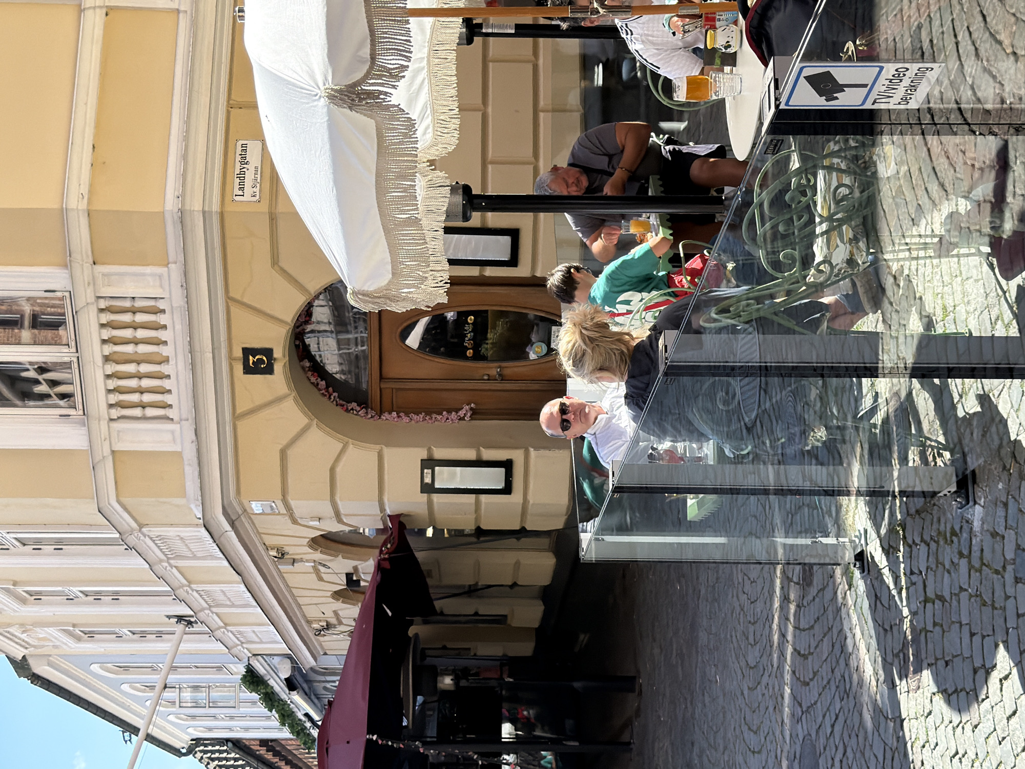 The image captures an outdoor seating area of a cafe or restaurant on a sunny day in Malmö, Sweden. The cafe is situated on a corner of a street with cobblestone pavement. The building facade is painted in a light yellow color with white architectural details, including decorative balconies and window frames. A street sign reads "Landbygatan" and "Kv. Stjärnan."
In the foreground, patrons are seated at tables. A glass partition encloses part of the seating area, reflecting the surrounding environment. Several individuals are visible: a man in sunglasses wearing a white shirt, a woman with blonde hair, a young person in a green t-shirt, and an older man in a grey t-shirt. They appear to be dining or socializing. One person is holding a drink with orange liquid, possibly juice. Another person is wearing a white shirt with black stripes.
Other elements include a large white umbrella with fringed edges, dark red awnings over other seating areas, outdoor heaters, and decorative greenery along the building. A sign with a CCTV camera icon and text "TV/video bevakning" is attached to the glass partition, indicating surveillance. The overall scene suggests a relaxed, public outdoor dining experience.