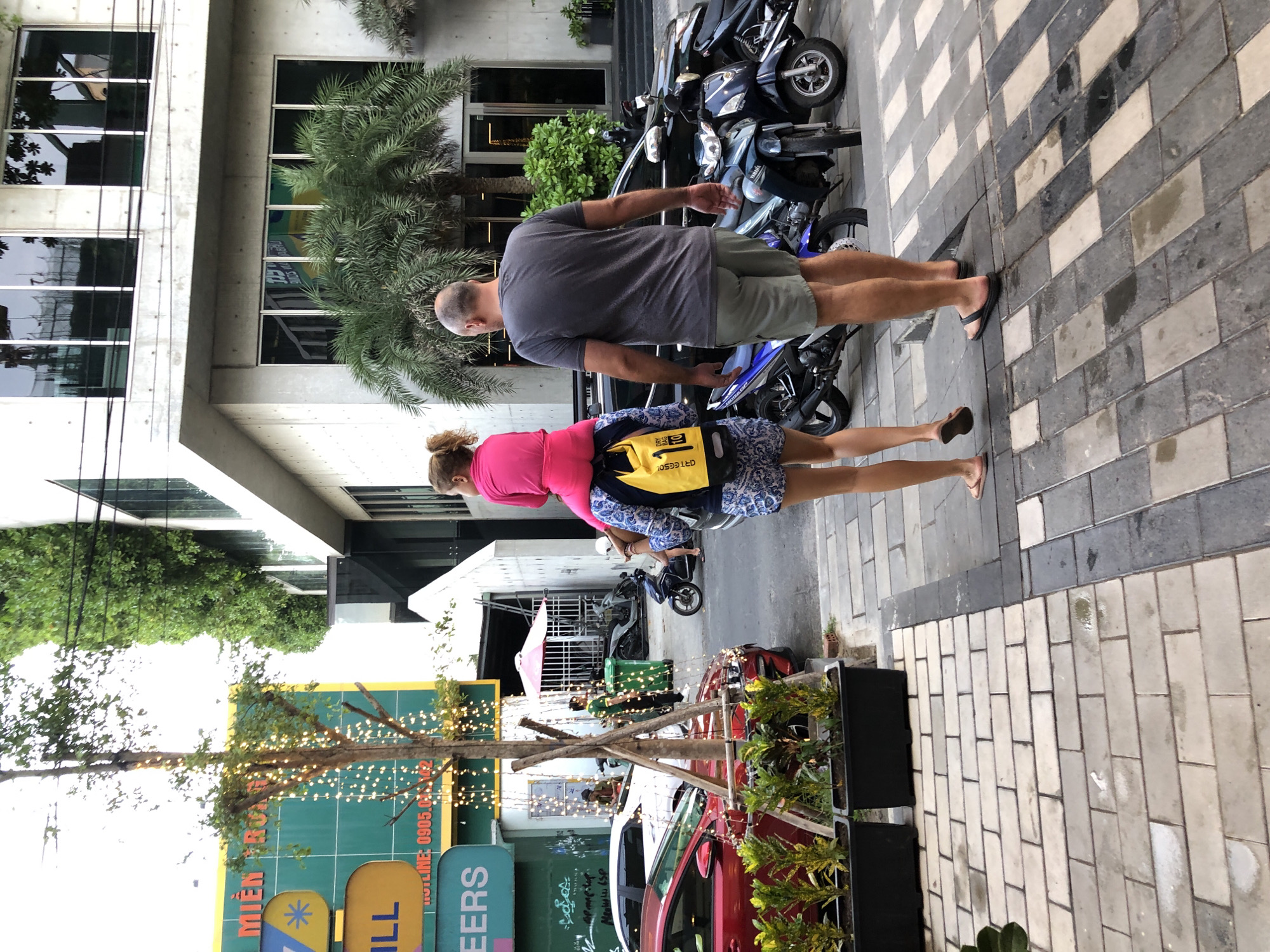 The image depicts two people walking down a street in Đà Nẵng, Vietnam. The man, wearing a grey t-shirt and olive shorts, walks ahead. Behind him, a woman in a bright pink top and patterned shorts carries a yellow backpack. They are walking on a sidewalk paved with light grey and white rectangular tiles.
To their left, there is a modern building with large glass windows and concrete architectural elements. Greenery, including palm trees, is visible near the building. Parked motorcycles are lined up on the street next to the sidewalk, suggesting a common mode of transportation in the area.
On the right side of the image, beyond the sidewalk, there are potted plants and what appear to be parts of vehicles, including a red car, hinting at a commercial or residential area with some businesses. The lighting suggests it is daytime, and the weather appears to be clear.
Visible text includes "MIỀN TRUNG" in large blue letters on a green background, and "HOTLINE 0905.043.342" in smaller white letters. There are also other signs with text and shapes, including a yellow sign with the word "ILL" and a blue sign with "ERS". The scene conveys a sense of everyday urban life in a Vietnamese city.