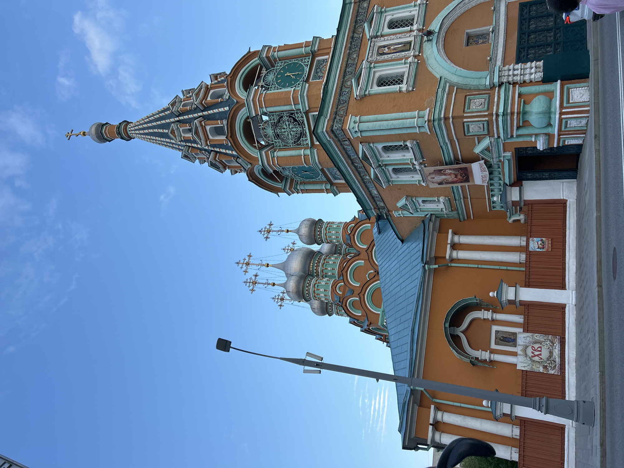 A low-angle view captures the detailed architecture of a building against a clear blue sky. The building features an orange facade with decorative elements in shades of green and white. It has multiple domes and a tower with a spire topped by a cross. The architectural style is reminiscent of Russian Orthodox churches. A streetlight with a simple lamp fixture is visible in the foreground, partially obscuring the building.