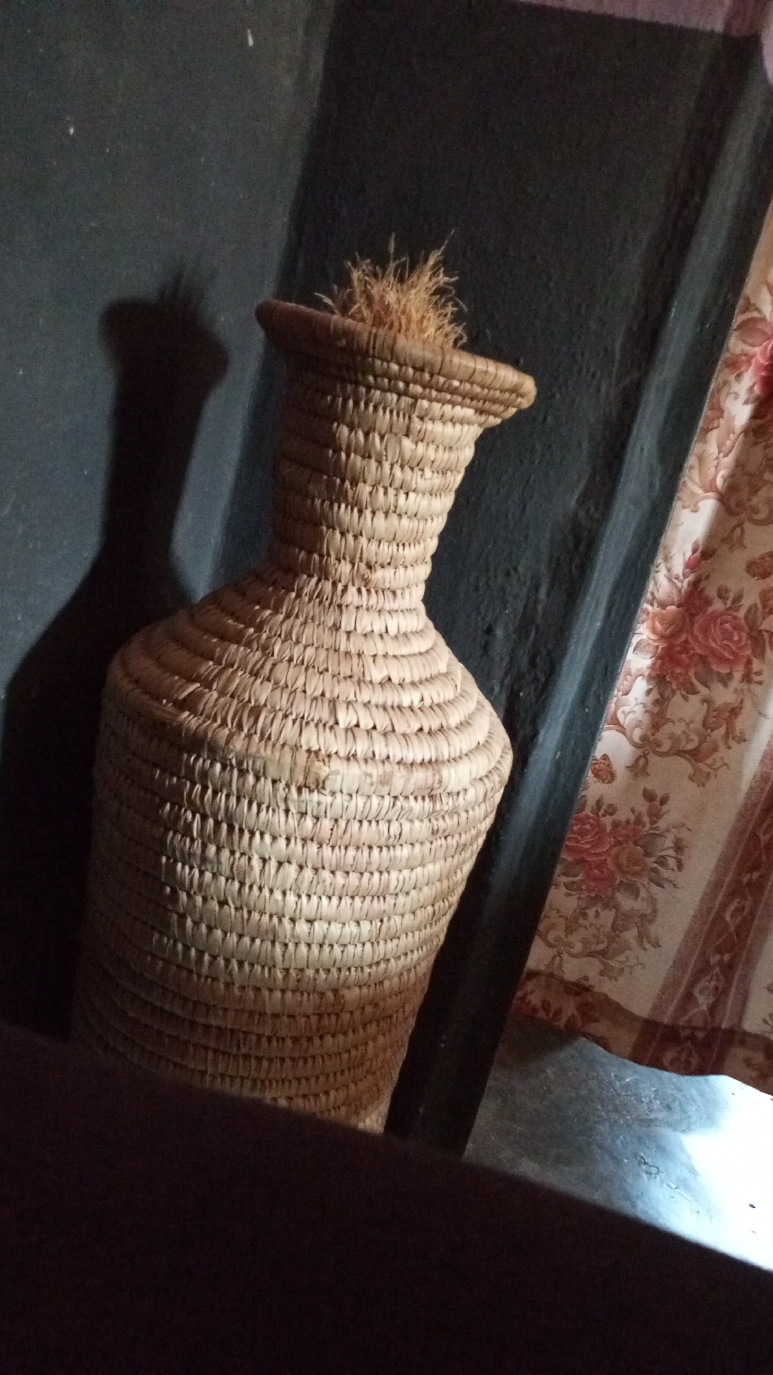 A woven vase sits in the corner of a room. The vase is tan with a narrow neck and wide body. It sits on a dark surface, likely a shelf or floor, in the city of Unknown, Unknown. Behind the vase is a dark wall and a curtain with a floral pattern. The vase casts a shadow on the wall.