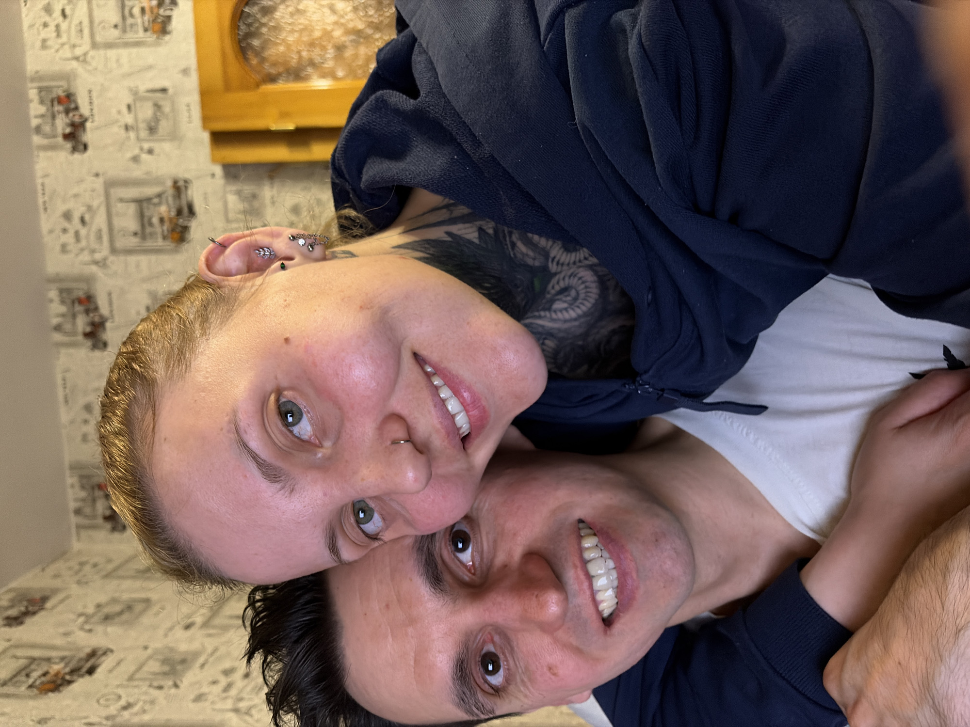 A man and a woman are posing closely for a picture. The woman is on top, with a visible tattoo on her neck, multiple ear piercings, and a nose ring. She is wearing a dark blue hoodie. The man is on the bottom and is also wearing a dark blue hoodie, and a white shirt is visible underneath. They are smiling and looking at the camera. The background features a patterned wallpaper with illustrations of Parisian landmarks. A wooden frame is visible on the wall above them. This image has been taken in a residential setting. It is not possible to confirm the location in Chaikovsky, Russia with certainty based on the given image.