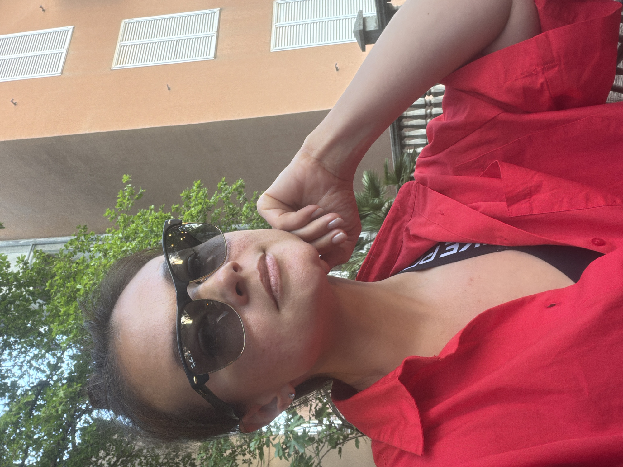 A woman is depicted in a close-up, half-body portrait, with her head tilted to her right and her gaze directed slightly upwards, obscured by dark sunglasses. She is wearing a vibrant red collared shirt, unbuttoned to reveal a black bralette or sports bra with white lettering on the band. Her left arm is bent at the elbow, and her hand rests near the side of her head. Her skin appears fair, and she has dark hair.
The setting is outdoors on a bright, sunny day. In the background, a building with a light orange or peach-colored facade is visible, featuring two prominent white louvered windows or shutters. Below these, a darker, shadowed architectural section extends across the building's width. Lush green foliage, including trees and bushes, is interspersed around the subject and in the mid-ground, suggesting a natural and urban environment. The general architecture and sunny disposition align with the location context of Budva, Montenegro.
Notable details include the "Ray-Ban" brand visible on the lens of her sunglasses. On the white band of her black bra, partially visible text reads "...KEP". The woman's expression, though hidden by sunglasses, appears calm and relaxed.
