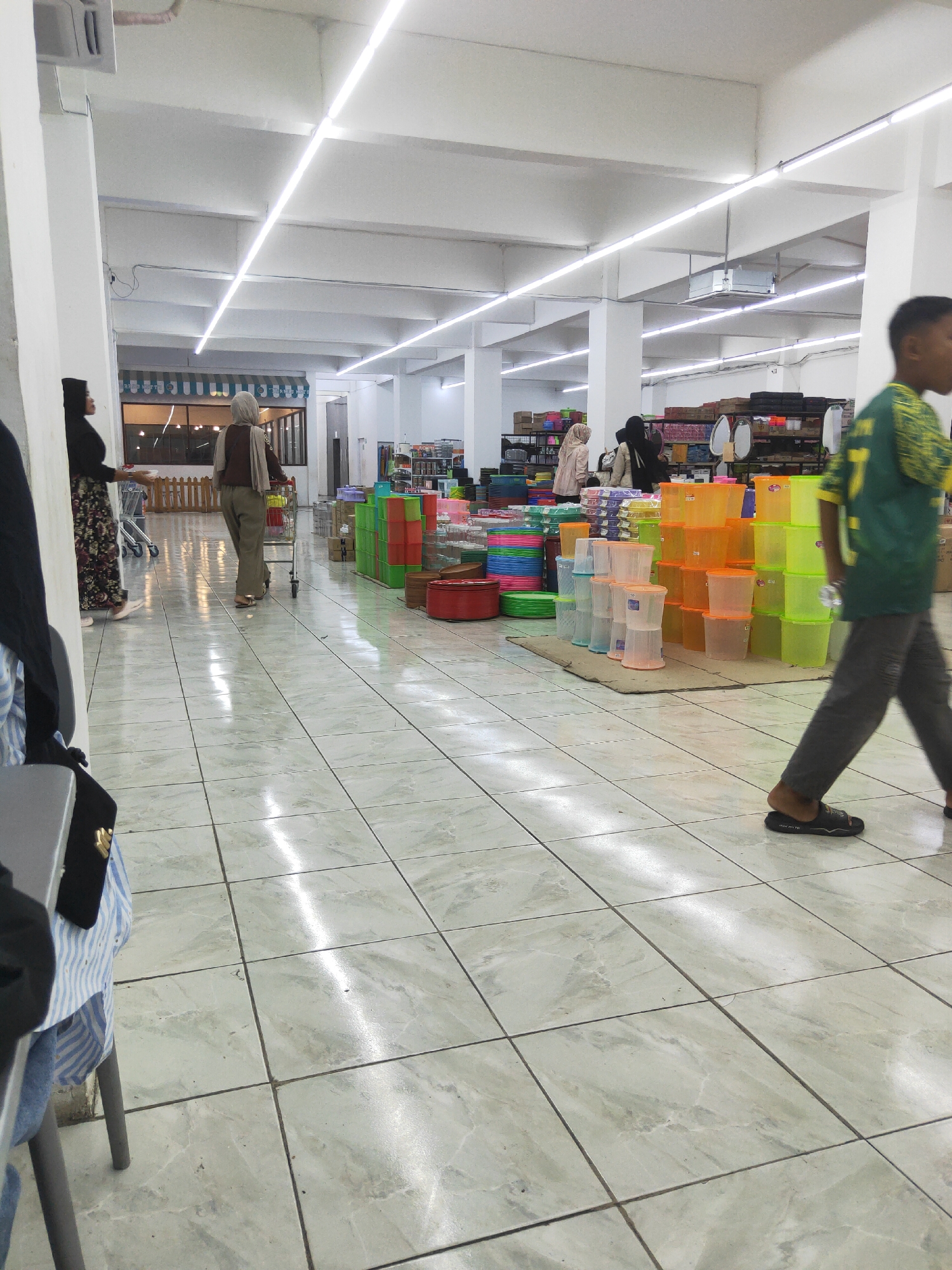 The image depicts the interior of a retail store, likely a supermarket or a housewares store, located in Sintang, Indonesia. The store features brightly lit aisles with white tiled floors that reflect the overhead fluorescent lighting. In the foreground, a young male wearing a green and yellow shirt and gray pants walks away from the camera. To the left, a woman in a black hijab and patterned skirt stands near a shopping cart, interacting with another woman in a light brown hijab and trousers who is pushing a shopping cart. The aisles are stocked with various goods, prominently featuring stacks of colorful plastic containers, buckets, and other household items in shades of green, orange, and blue. Shelving units are visible in the background, displaying more merchandise. The overall impression is one of a busy commercial environment.