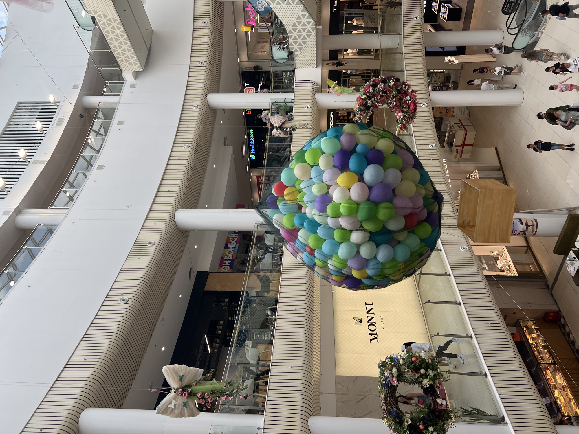 This indoor shot depicts a multi-story commercial center. It appears to be the interior of a modern shopping mall. The view looks upward from within the mall. A large, colorful display made of many balloons is suspended in the open space of the central atrium. Also suspended are decorative wreaths and a stylized dress form.
The mall's architecture is visible. Smooth white walls with horizontal ribbed detailing curve inward toward the central area. Large retail storefronts are visible on multiple levels. The storefronts have glass facades allowing views of displays and merchandise. Store signs such as "MONNI," "Health Store," and "ОН БОНБОН" are present. Some stores display sale signs of "-30%".
In the background, on the ground floor, people are walking through the mall. An escalator is partially visible.
The location is identified as the city of Plovdiv, Bulgaria.
