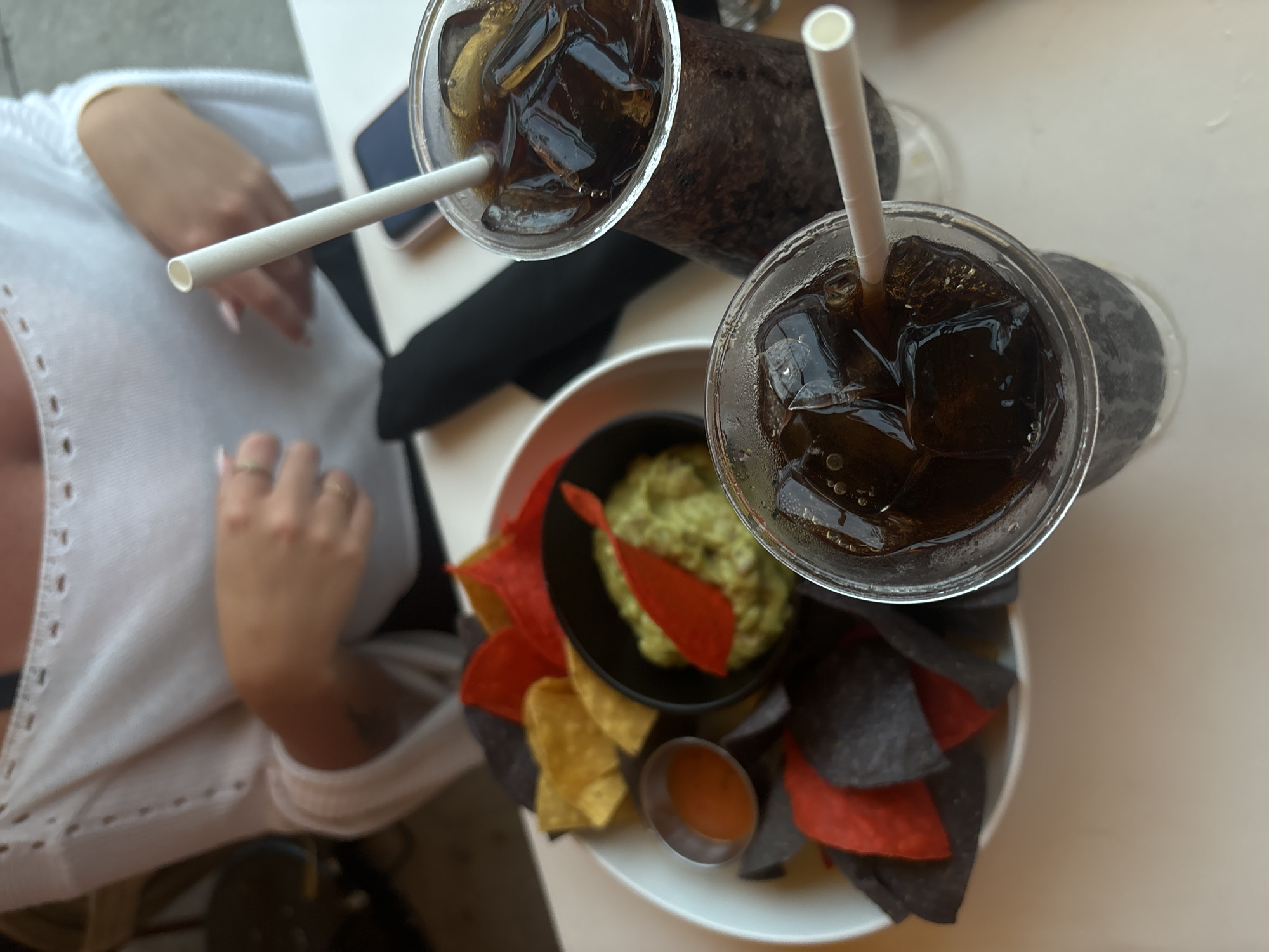 An overhead medium shot captures a table at an outdoor restaurant in Miami Beach, United States of America. In the foreground, two plastic cups filled with ice and a dark beverage, likely soda, are placed on a white table. Each cup contains a white paper straw. To the left, a white bowl holds a generous portion of guacamole in the center, surrounded by a variety of tortilla chips, including blue, yellow, and red varieties. A small silver dish containing a reddish sauce sits to the left of the chips. In the background, a person wearing a white, bell-sleeved top with cut-out detailing is partially visible, with their hands resting on the table. The overall scene suggests a casual dining experience.